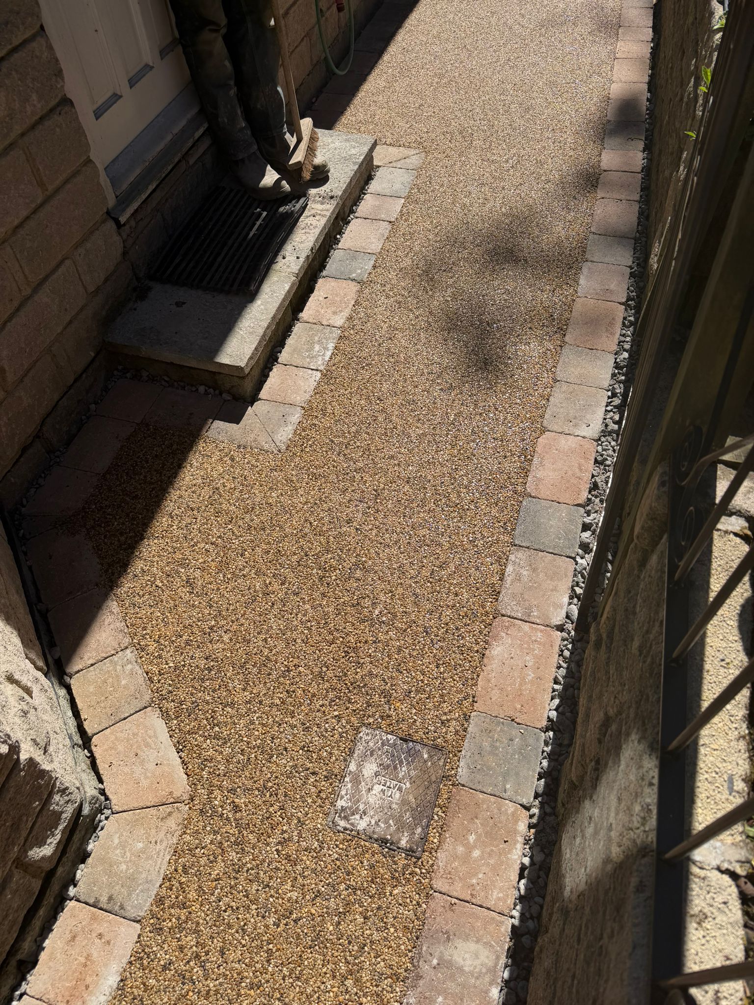 Narrow gravel walkway bordered by brick edges beside a building and fence in sunlight