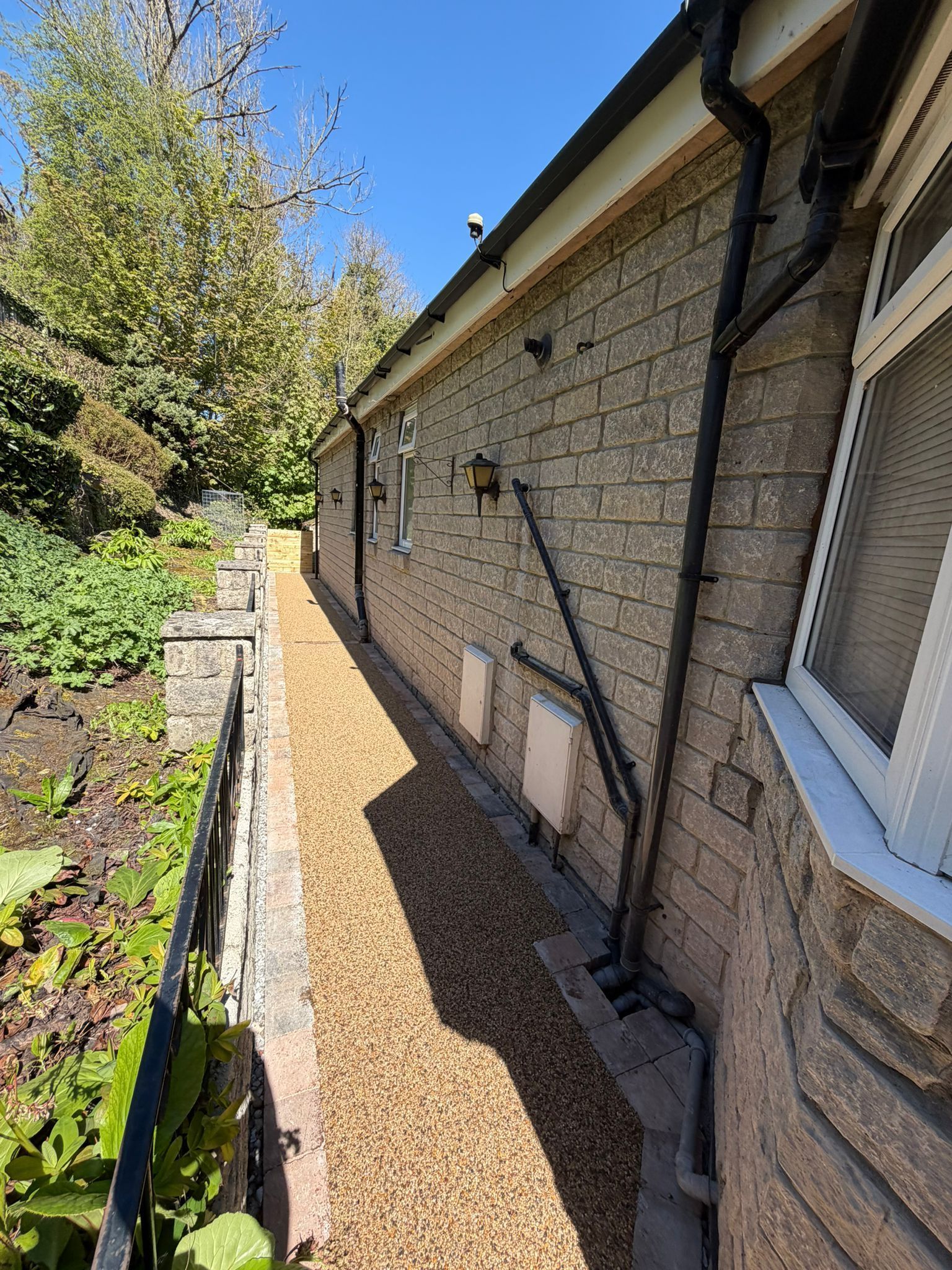 Narrow gravel path beside a stone house with windows, garden beds, and trees in bright sunlight