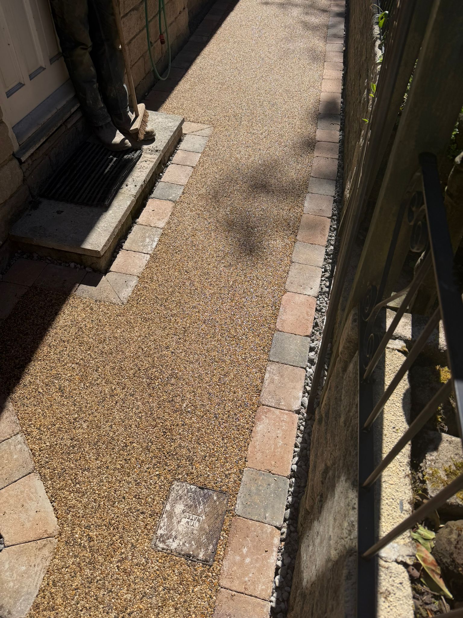 Narrow gravel path beside a building, bordered by brick edging and metal fencing.