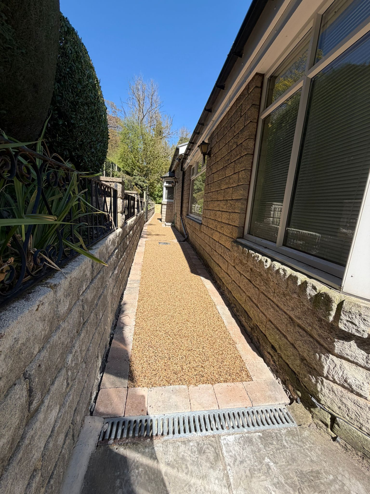Narrow gravel walkway beside a brick house and hedge, with windows and a drainage grate under a blue sky