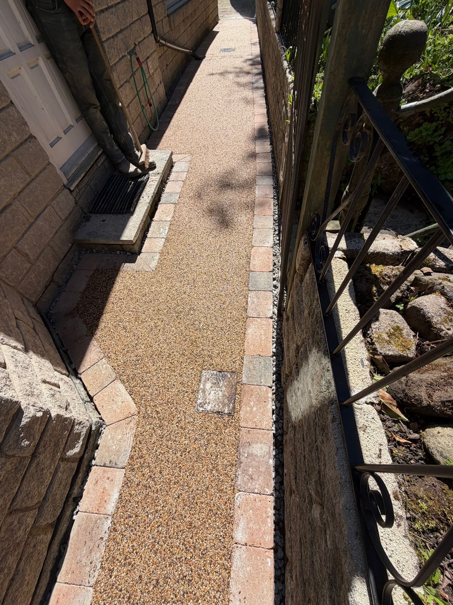 Narrow gravel walkway beside a brick house, with a black metal fence and shrubs on the right.