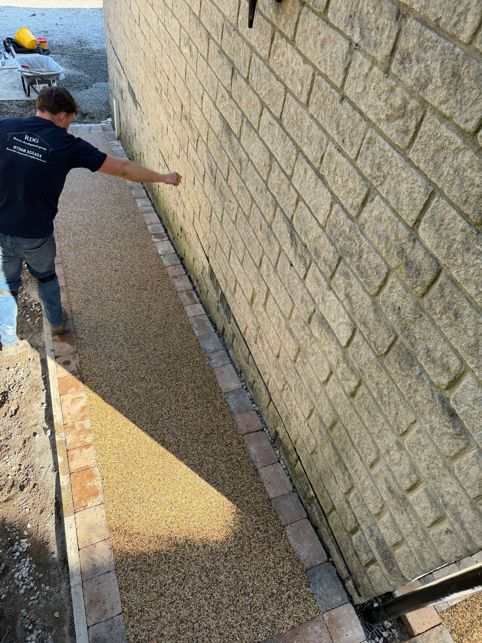 Person spreading gravel along a narrow strip beside a stone retaining wall.
