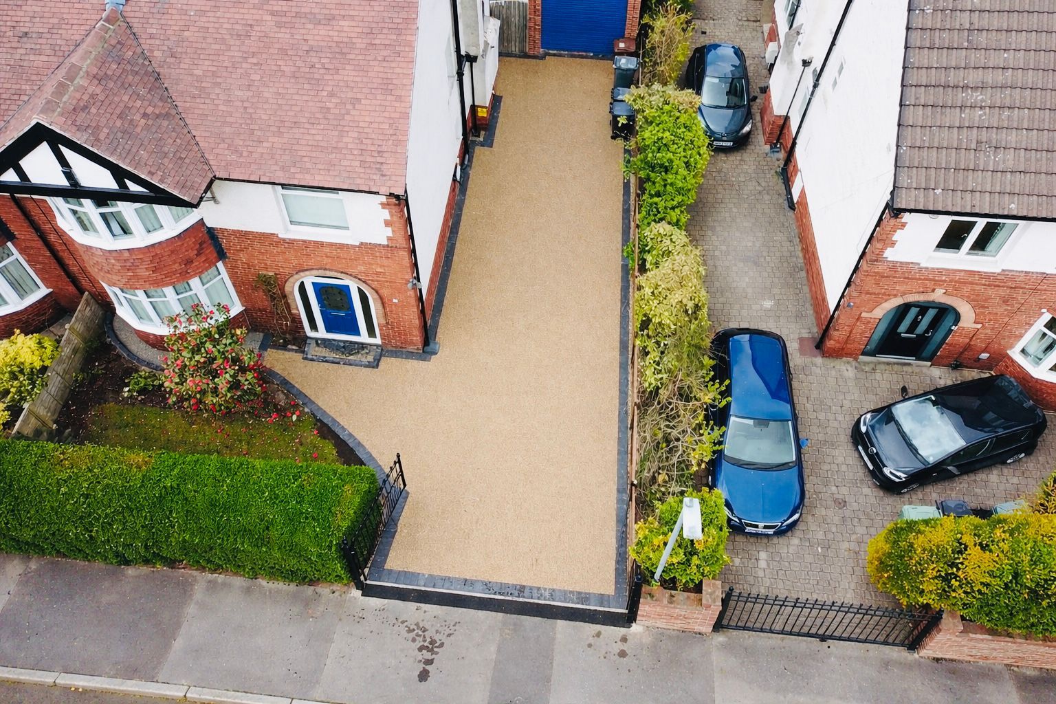 Aerial view of two houses with driveways, parked cars, and a paved front yard between brick homes