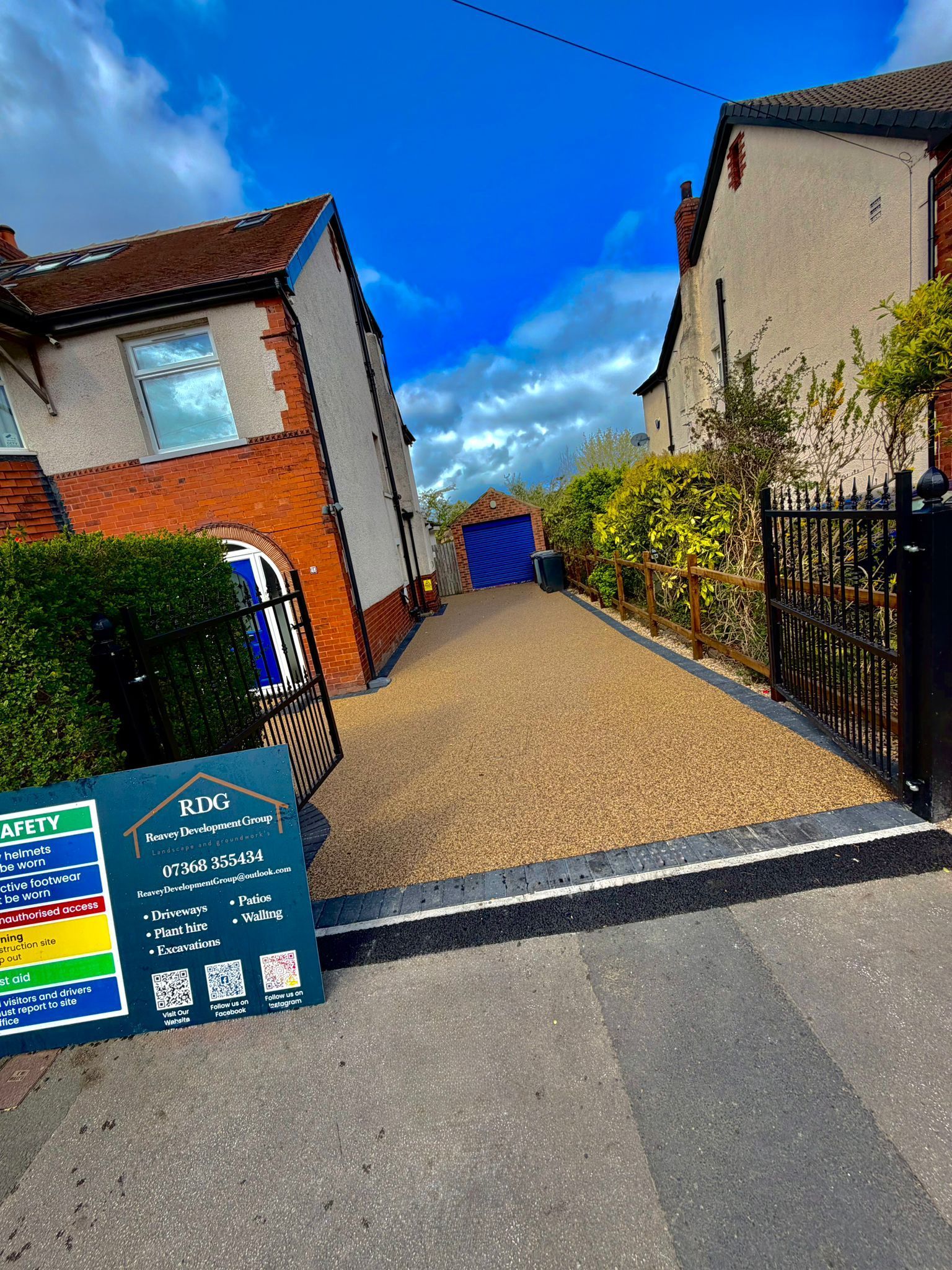 Residential driveway entrance with houses, hedges, and a blue sky; a signboard sits in the foreground.