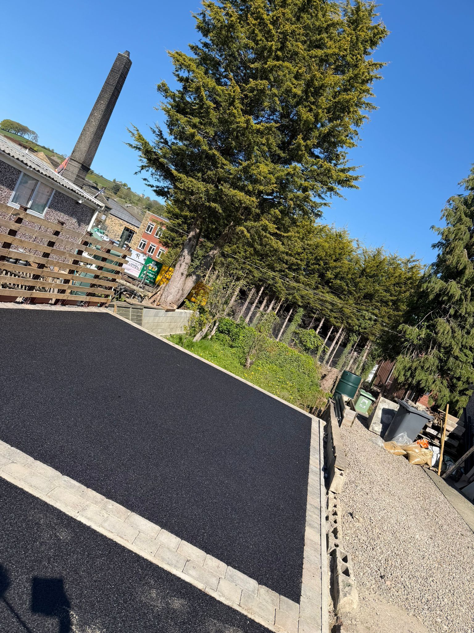 Outdoor patio with black paving, a large tree, and gravel path under a clear blue sky