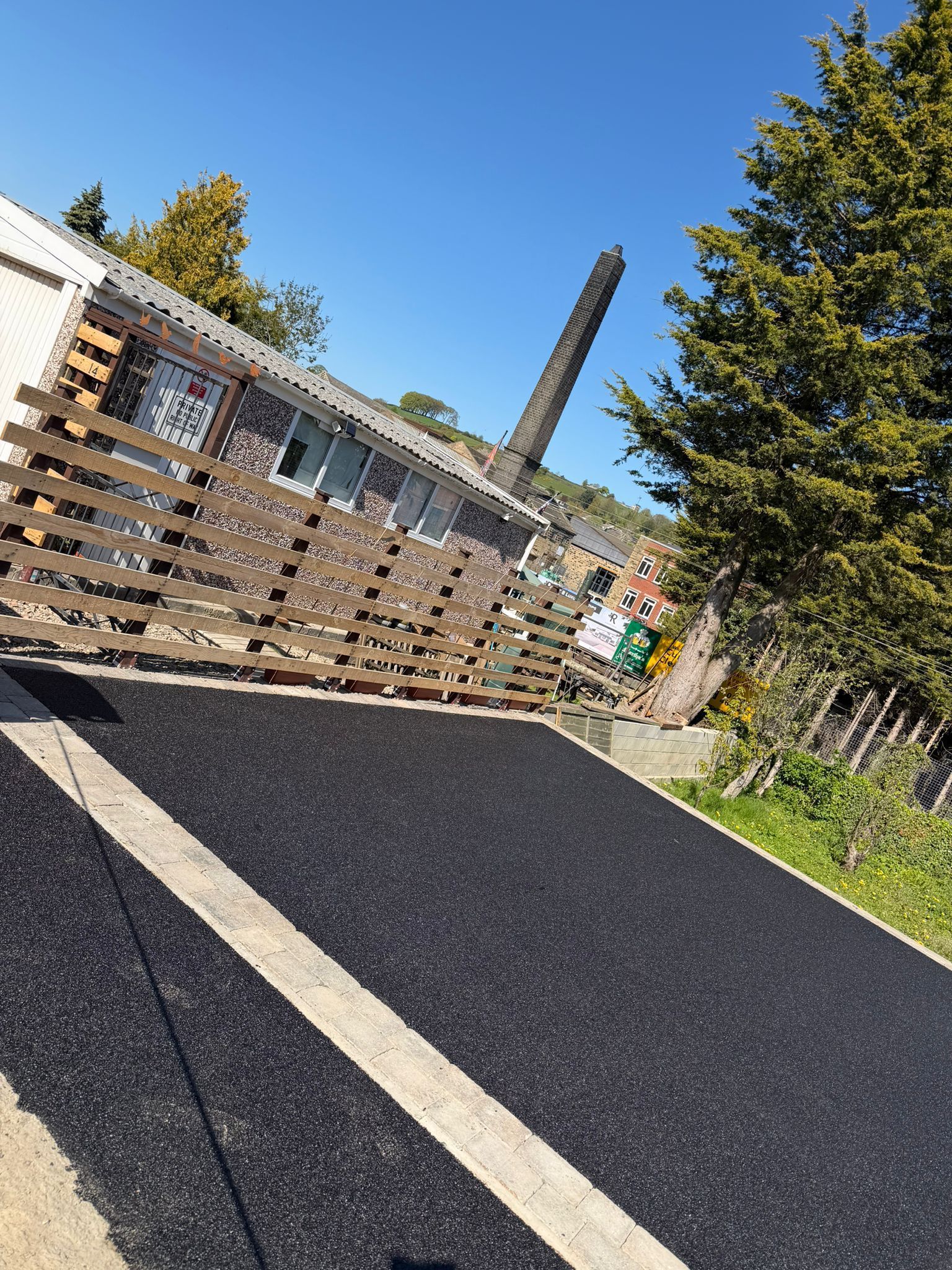 Tilted view of a black paved path beside a wooden fence, trees, and a building under a clear blue sky