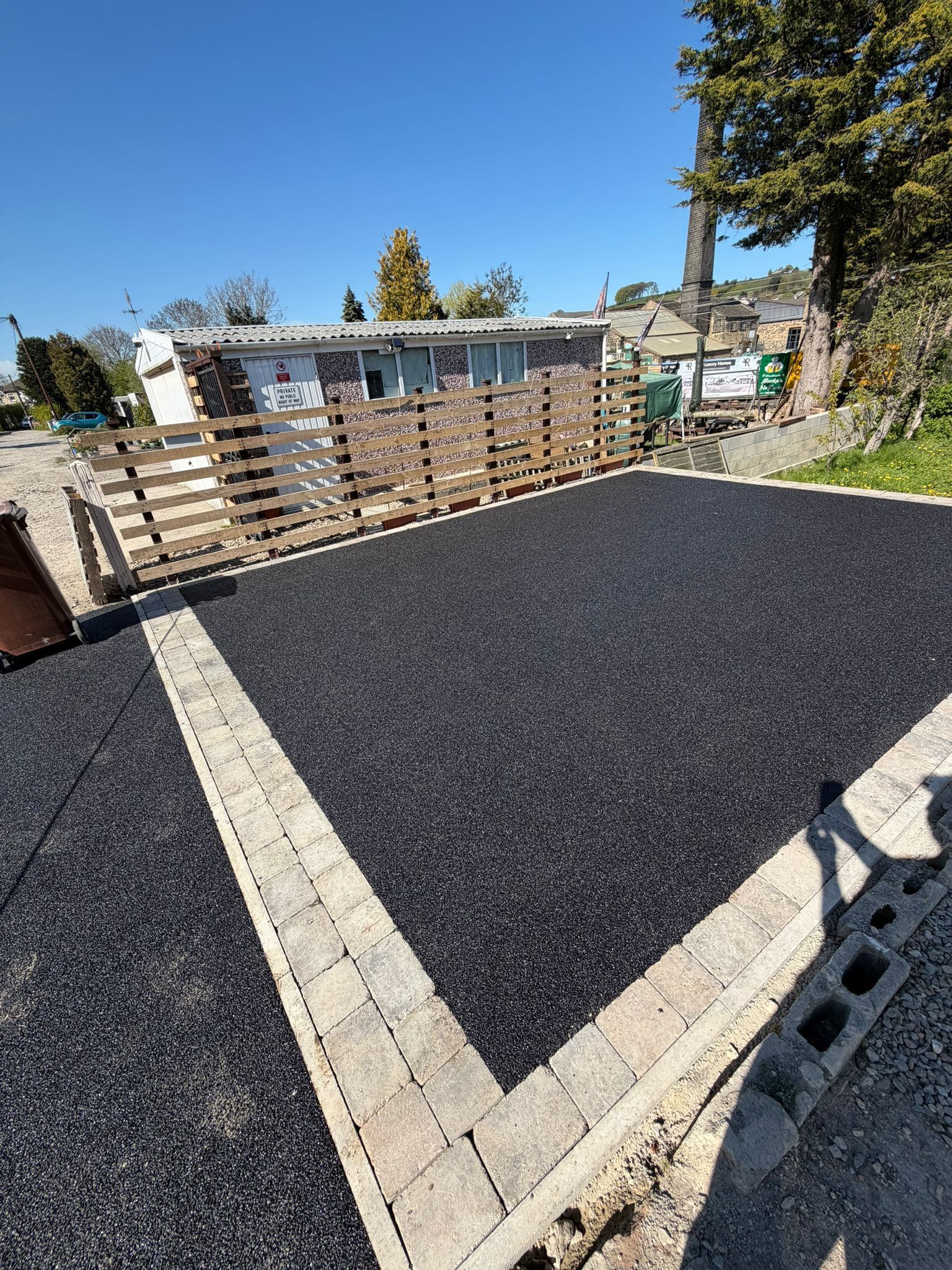 Freshly paved black asphalt driveway bordered by light stone pavers beside a wooden fence and house.