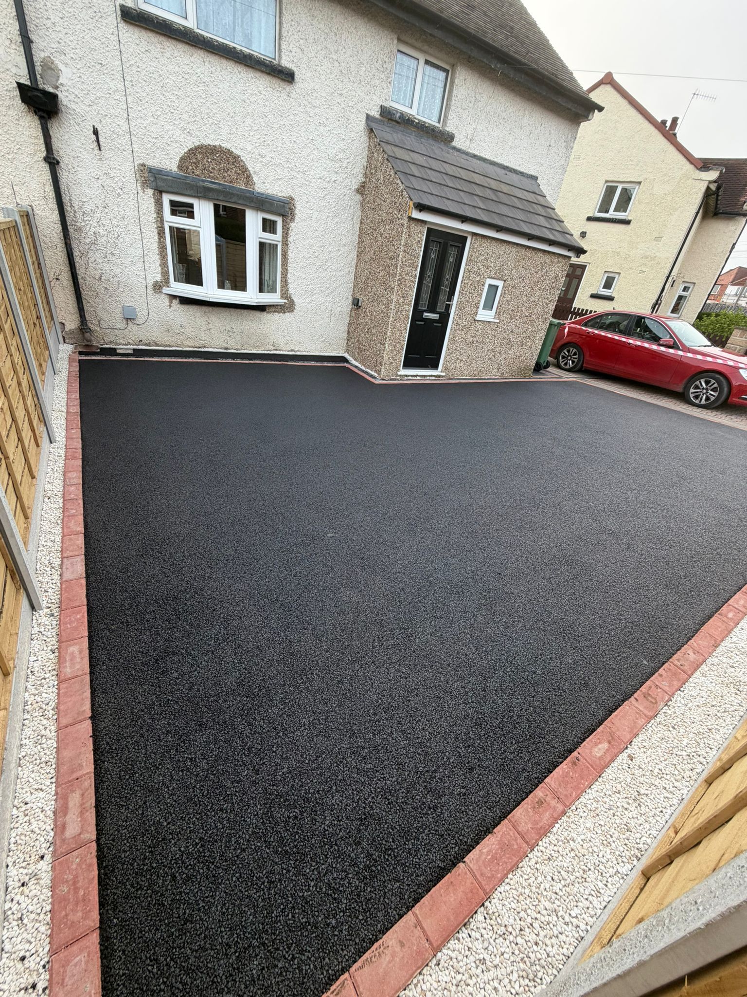 Fresh black asphalt driveway beside a stone house, with a red car parked on the street.