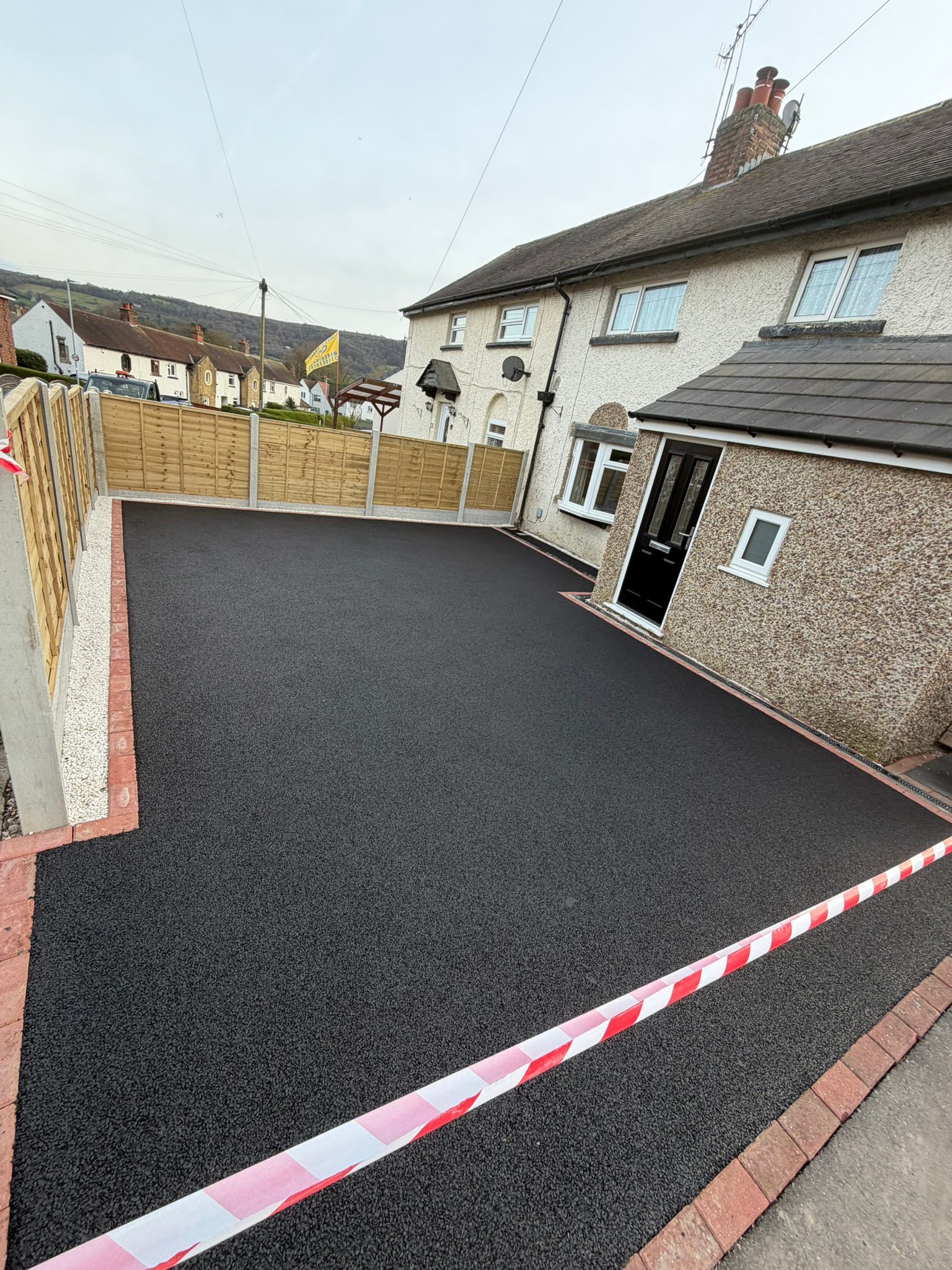 Freshly paved black asphalt driveway beside a house, bordered by brick edging and red-and-white tape