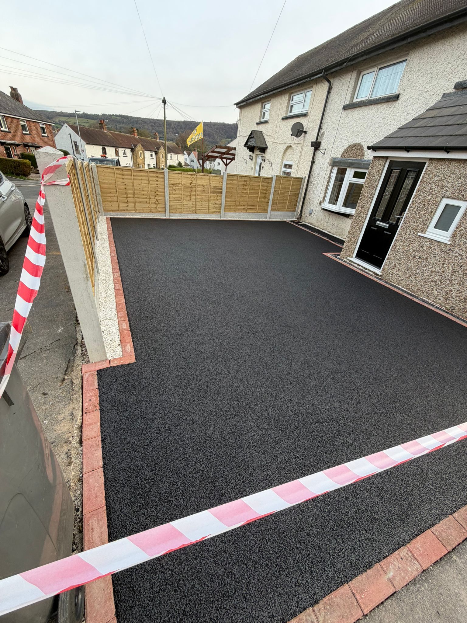 Freshly resurfaced black driveway beside a house, bordered by pink-and-white tape and fencing.