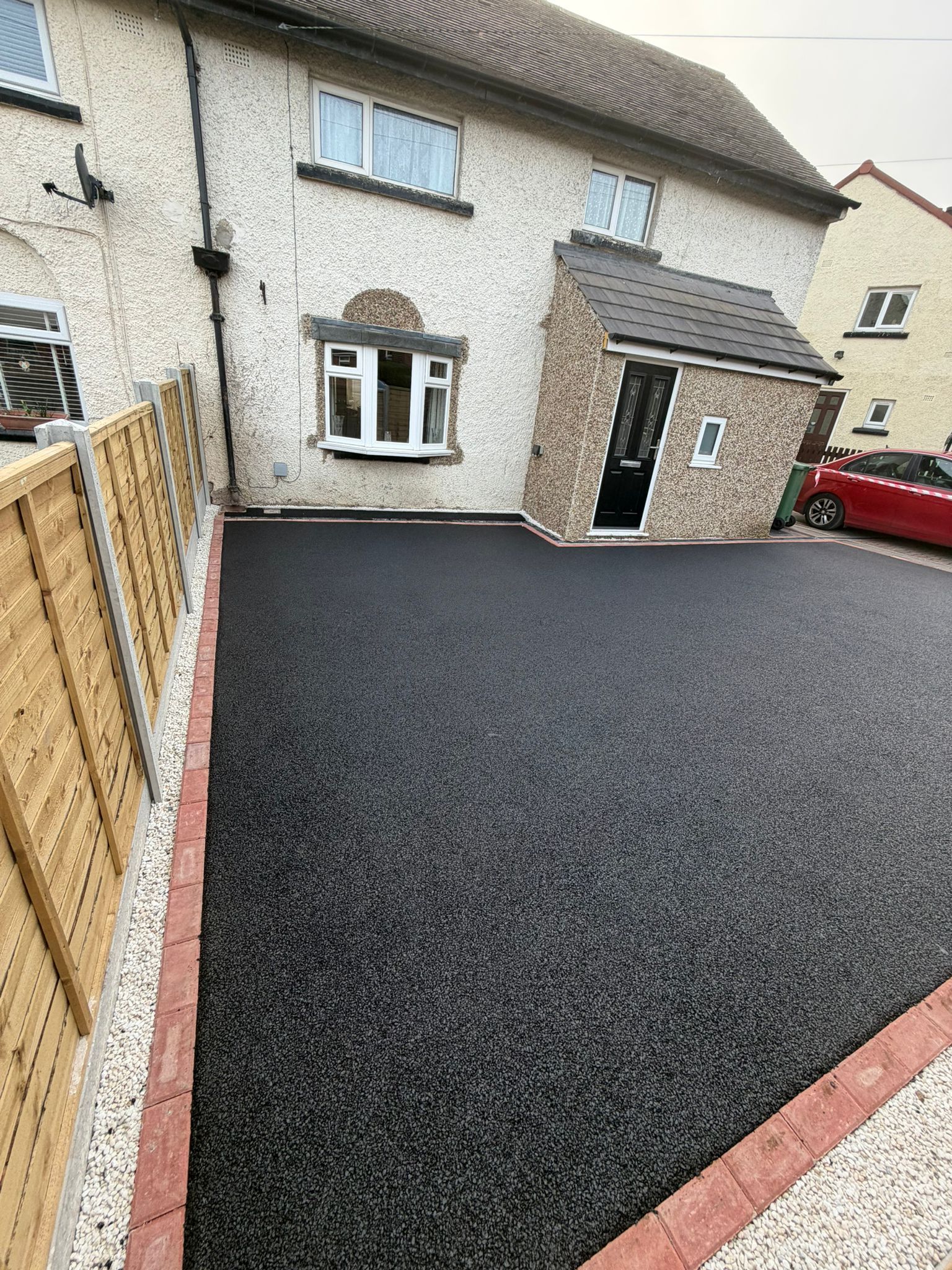 Freshly paved black driveway with red brick edging beside a cream house and wooden fence