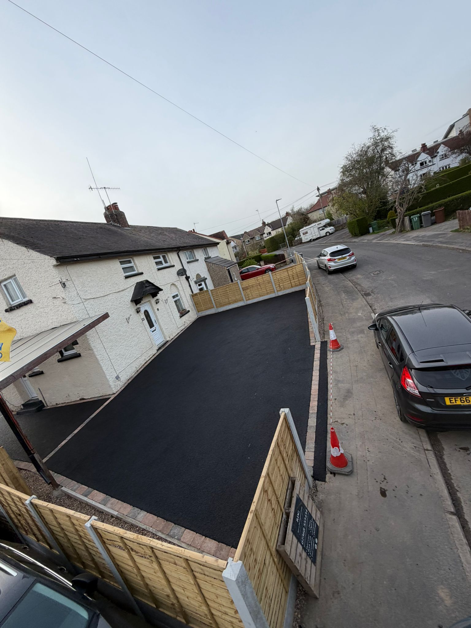 Tilted street view of a house, driveway, parked cars, and a road with traffic cones under a cloudy sky