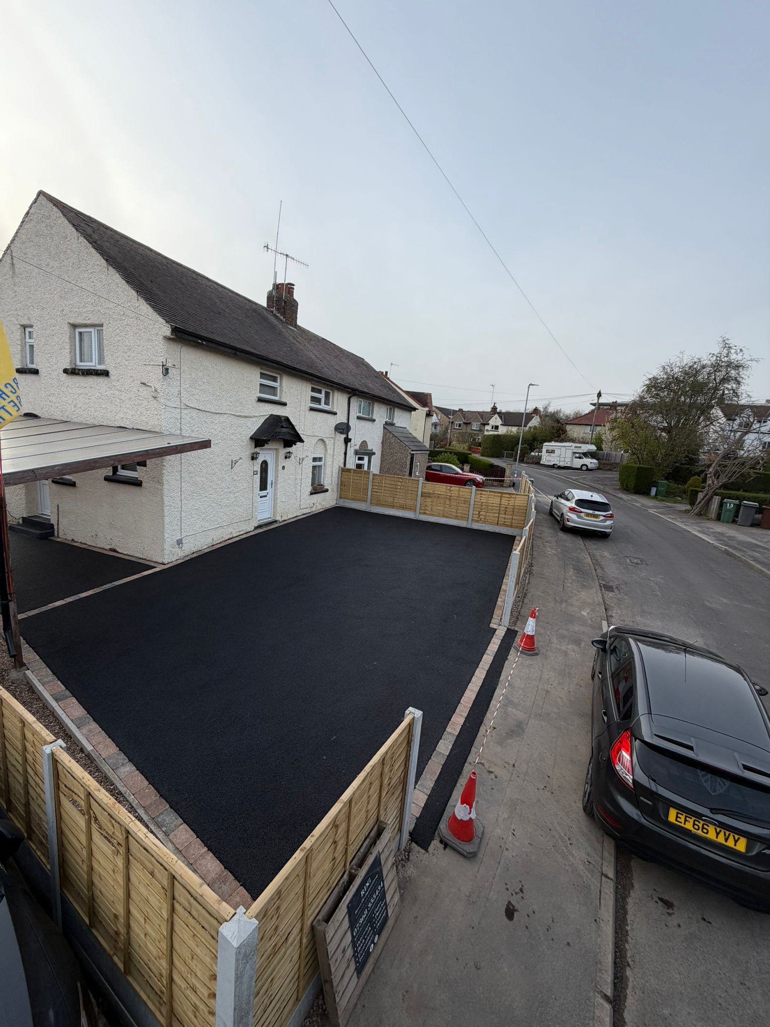 Residential street with a white house, black driveway, parked cars, and roadworks barriers nearby