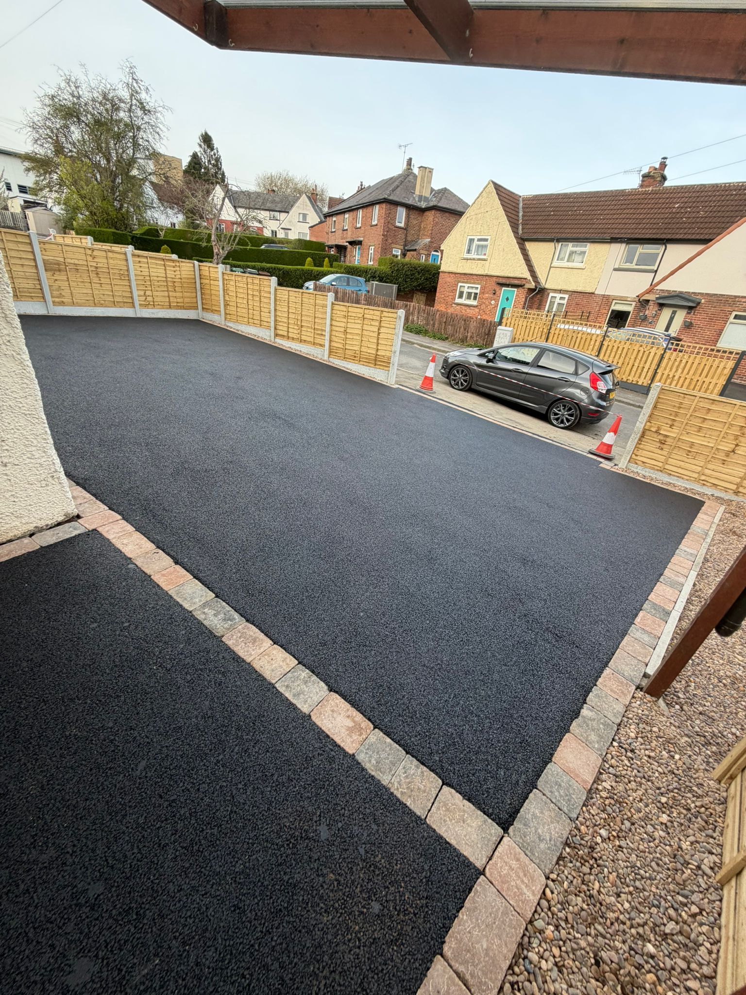 Freshly paved black driveway beside a house, bordered by low brick edging and yellow fencing.