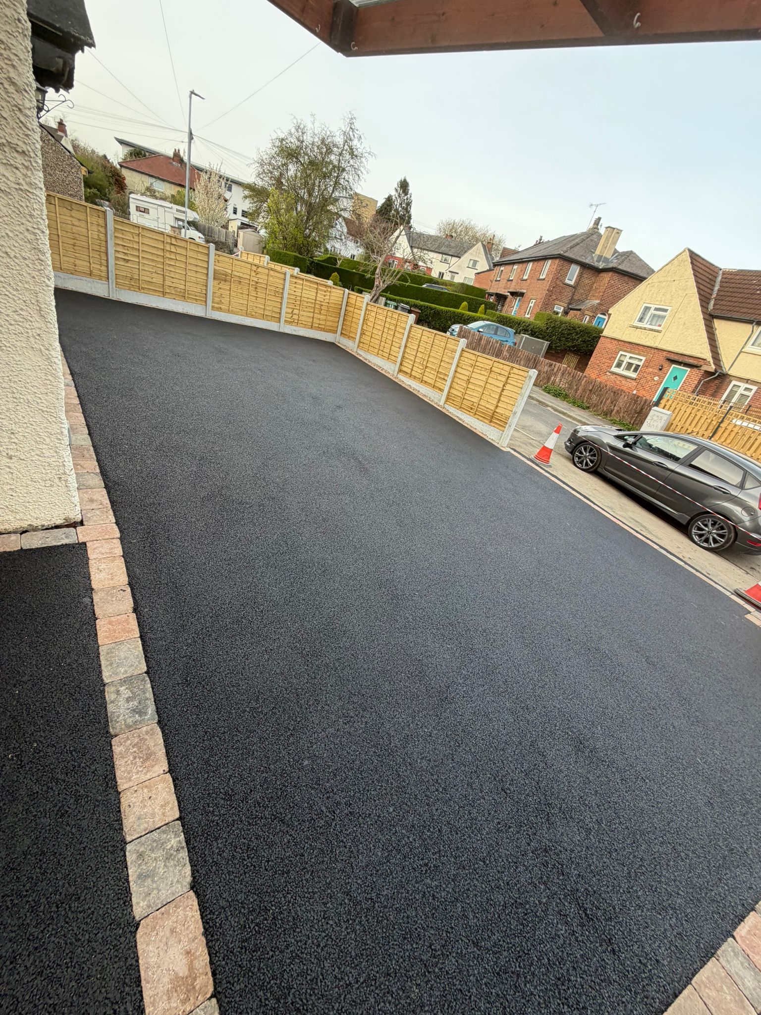 Freshly paved black driveway beside a house, with a fenced street and parked car beyond.