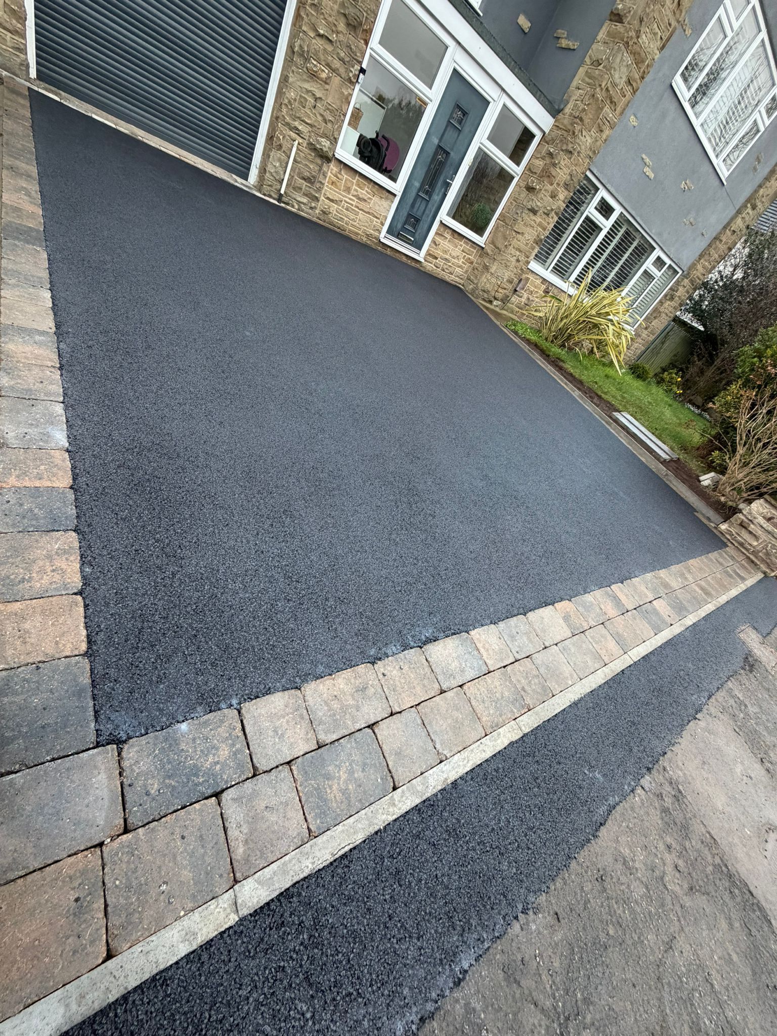 Paved driveway with dark resin surface, light stone border, and a brick house entrance in the background