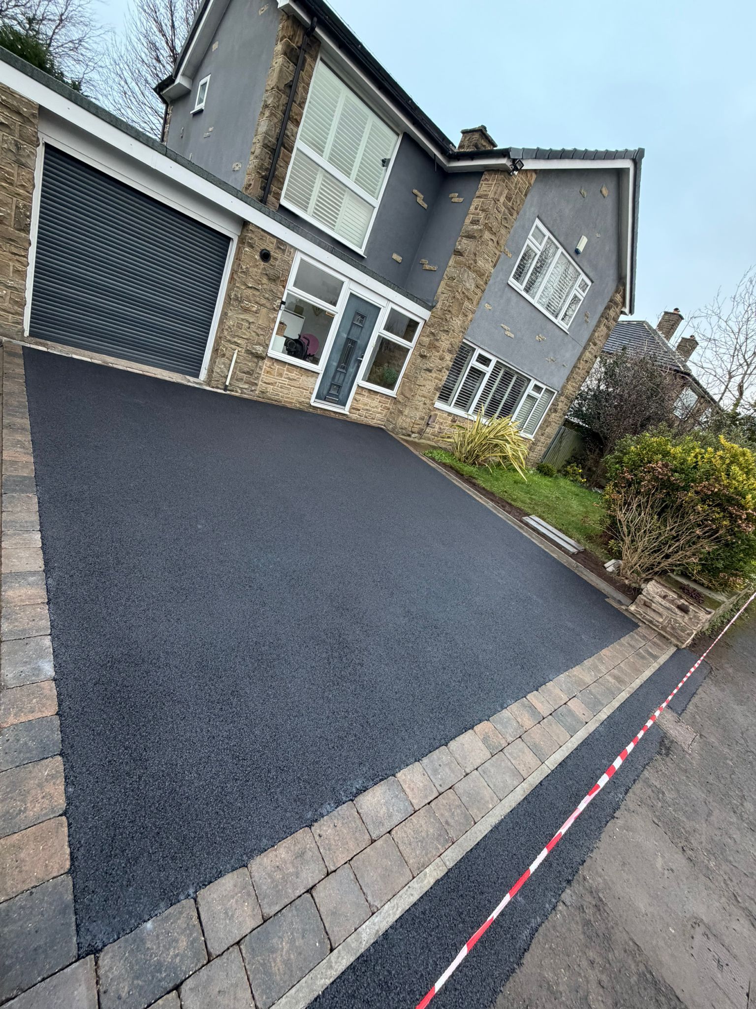 Driveway with fresh black asphalt leading to a gray stone house and garage, viewed at a tilted angle.