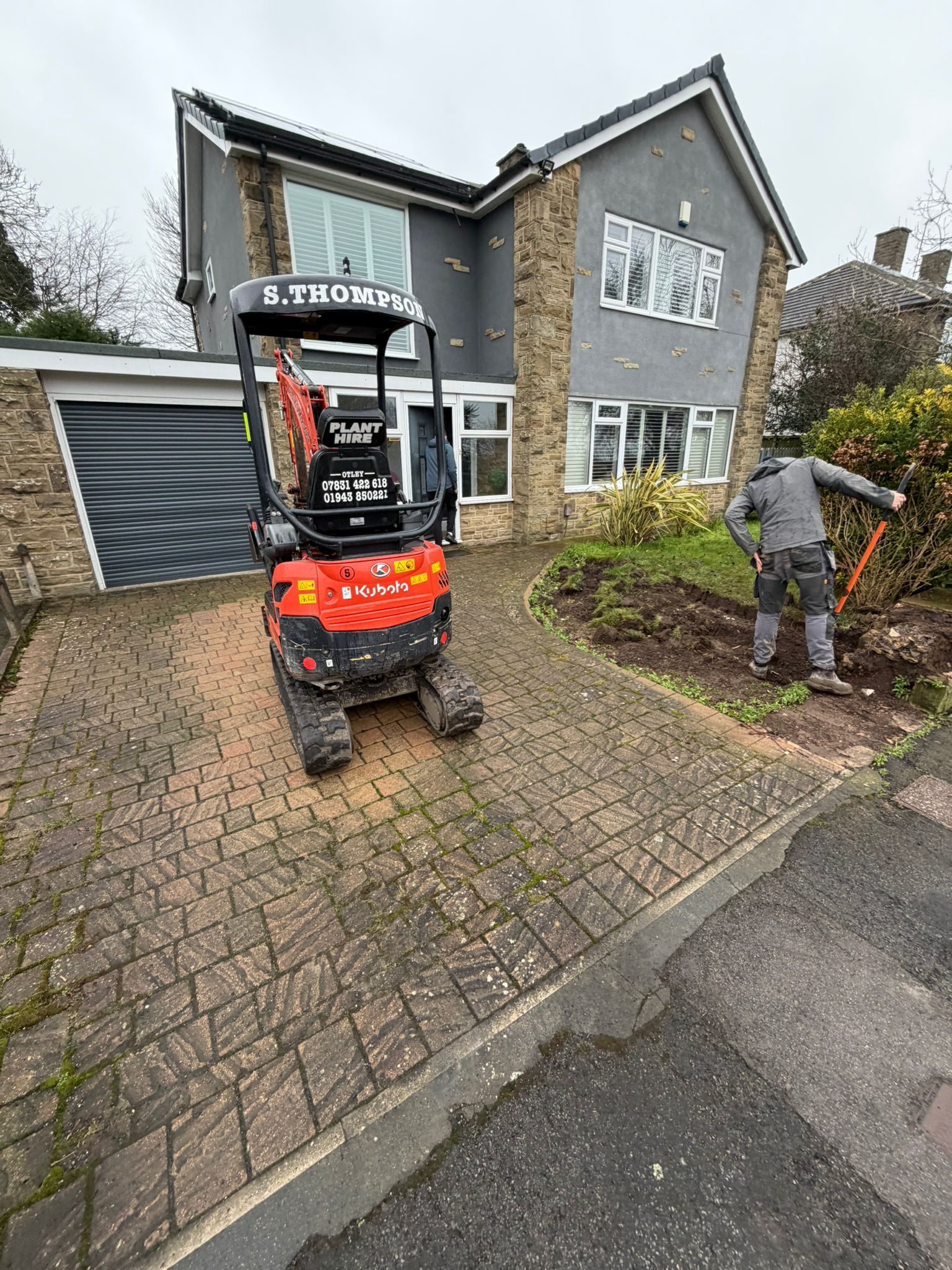 Mini excavator parked in a driveway beside a gray house, with a worker trimming the front garden.