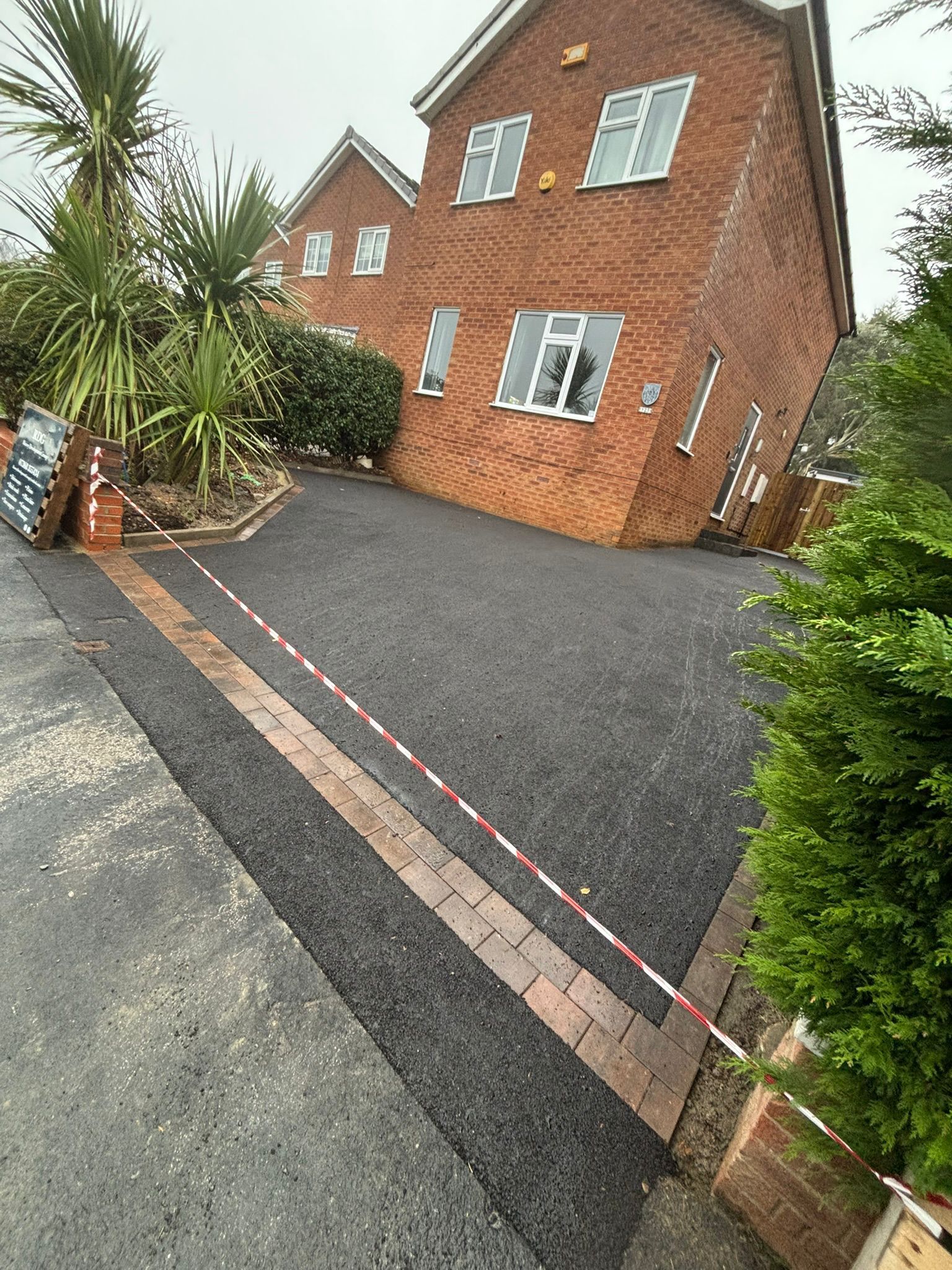 Tilted view of a red brick house with a black driveway, palm tree, and striped barrier tape across the foreground