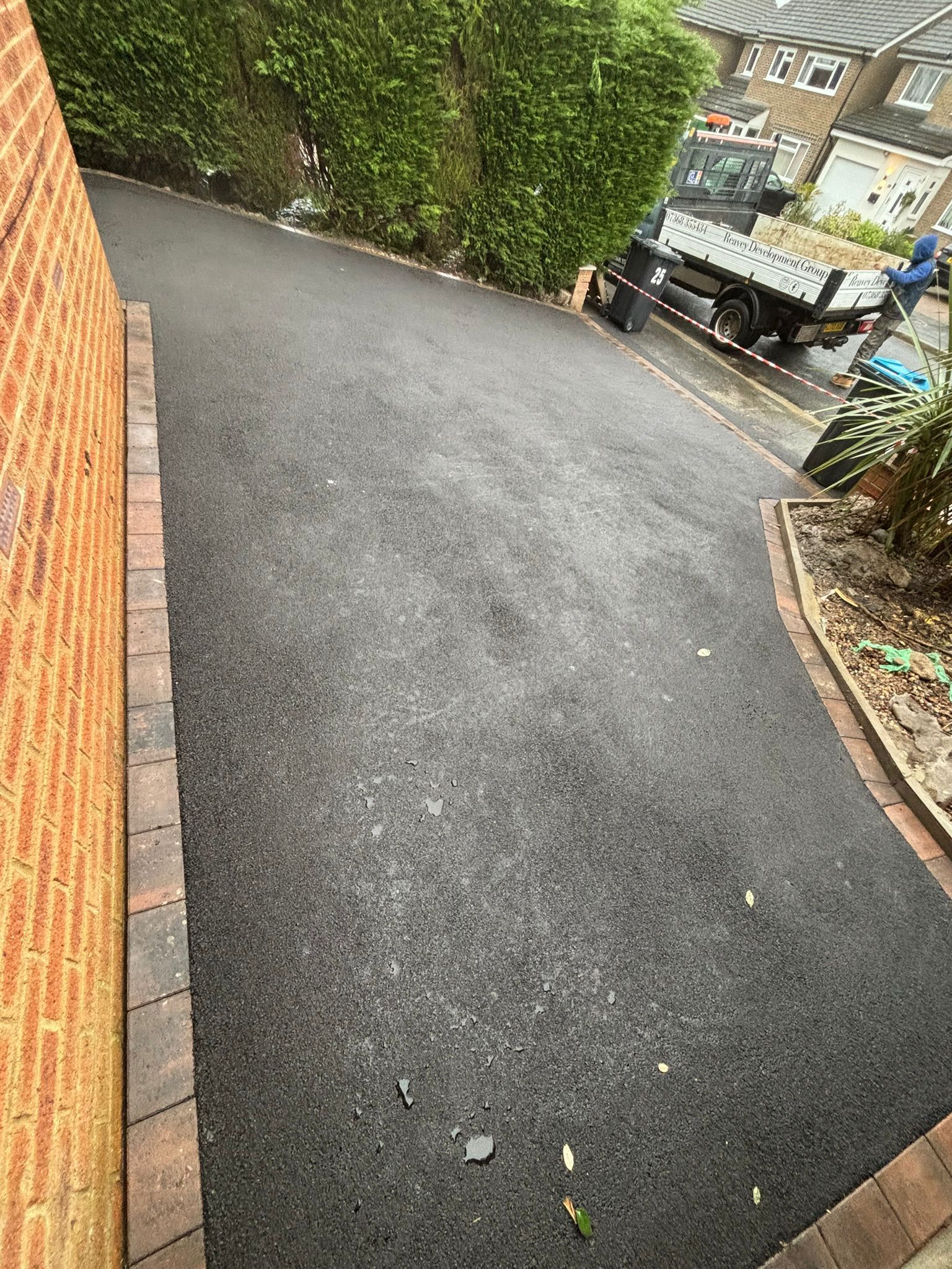 Narrow wet asphalt path beside a brick wall and hedge, leading downhill toward parked cars.