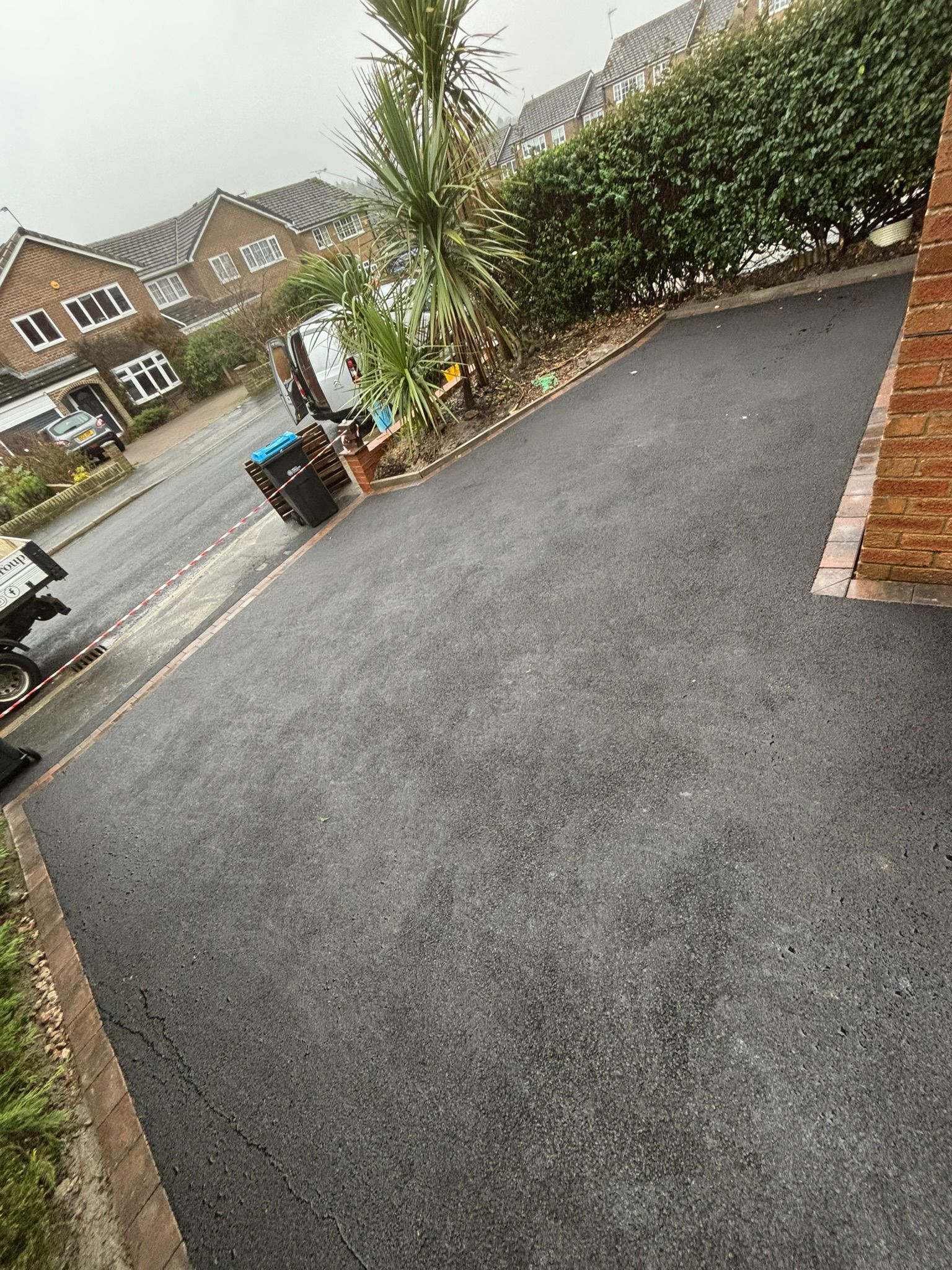 Residential driveway with fresh black asphalt, brick edging, and hedges beside a quiet street.