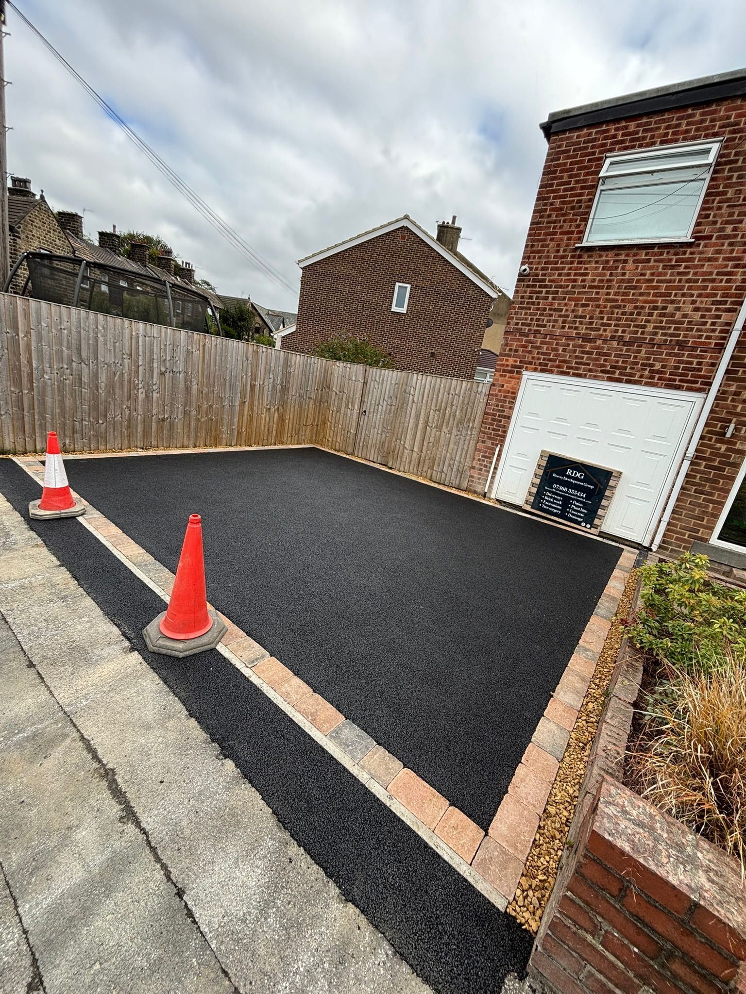 Newly paved driveway with orange cones beside a brick house and wooden fence