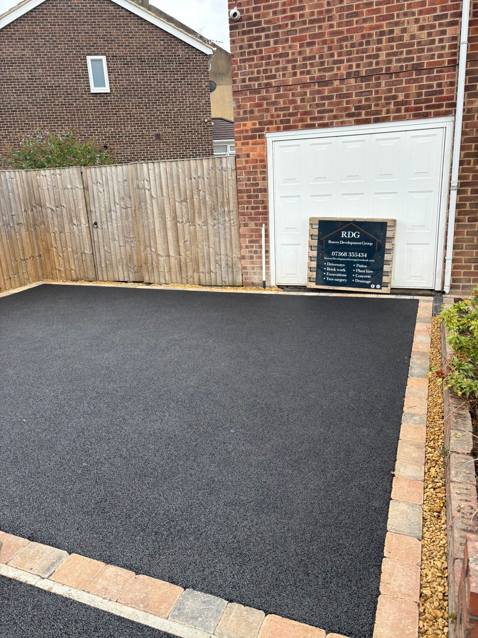 Freshly paved black driveway beside a brick house and white garage door, with a skip at the back.