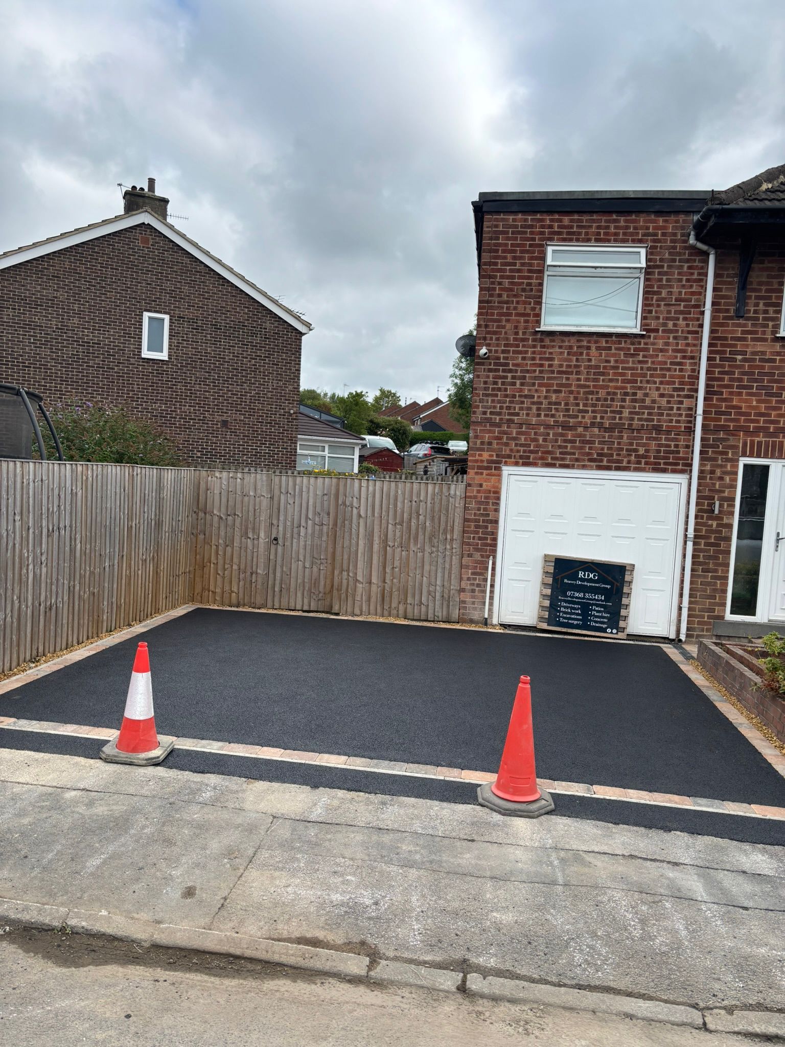 Empty paved parking space with two orange cones beside a brick house and wooden fence