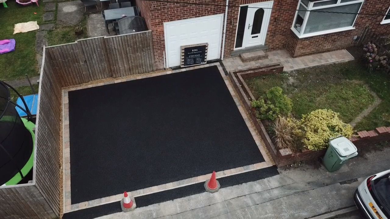 New black asphalt driveway beside a brick house, with two orange cones near the street