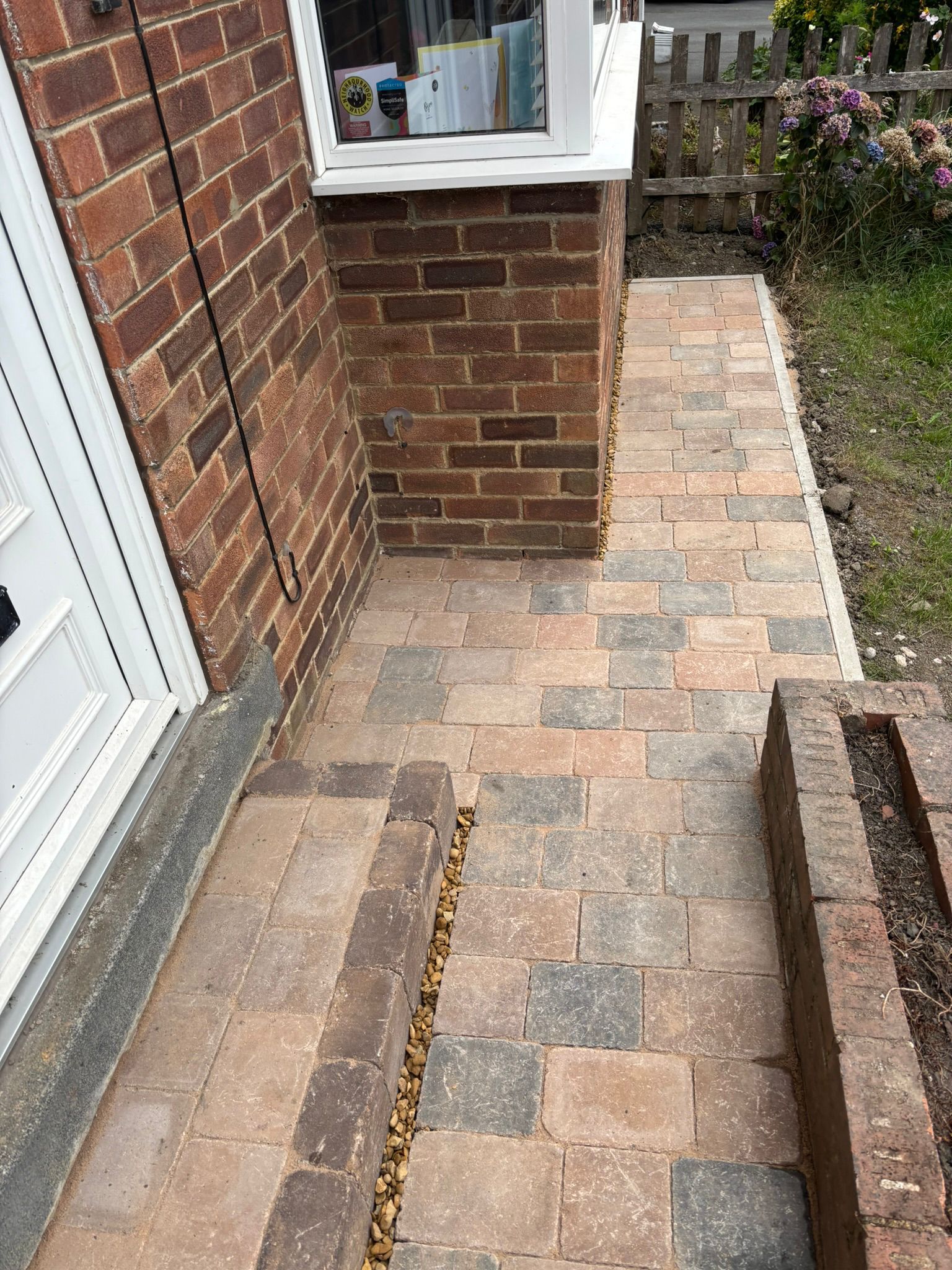 Brick patio walkway beside a house, with paved stones, garden edge, and a doorway.
