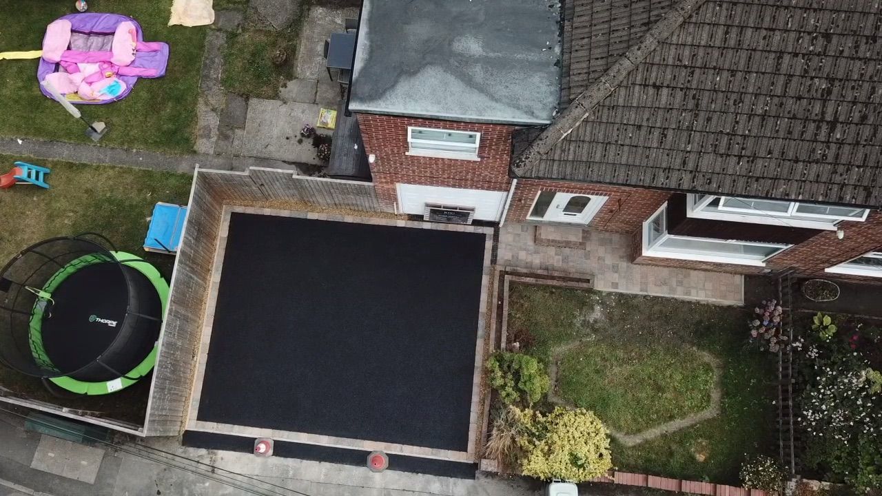Aerial view of a backyard with a trampoline, patio, lawn, and the roof and rear wall of a house.
