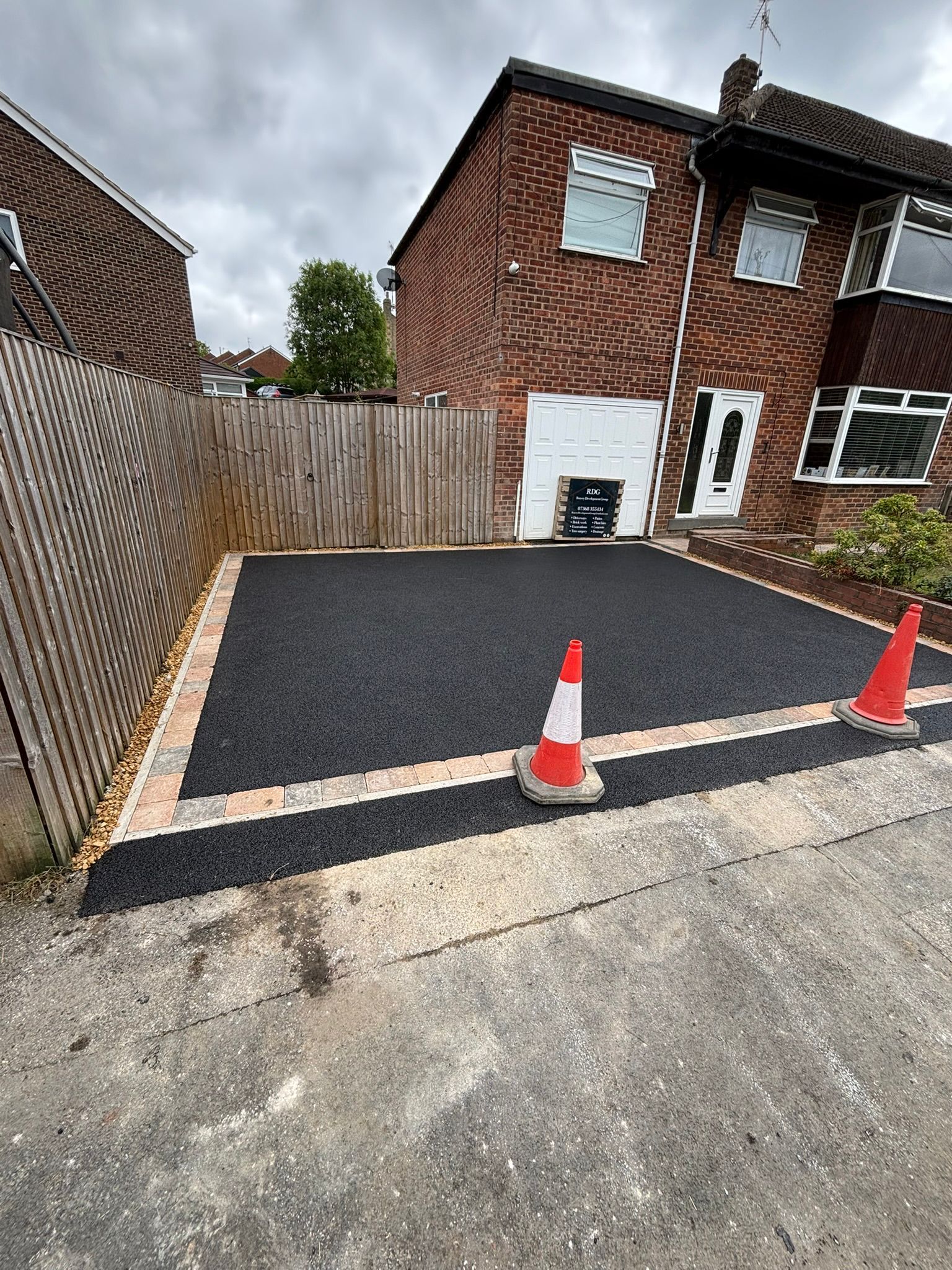 Fresh black asphalt driveway beside a brick house, with two orange cones and a wooden fence.