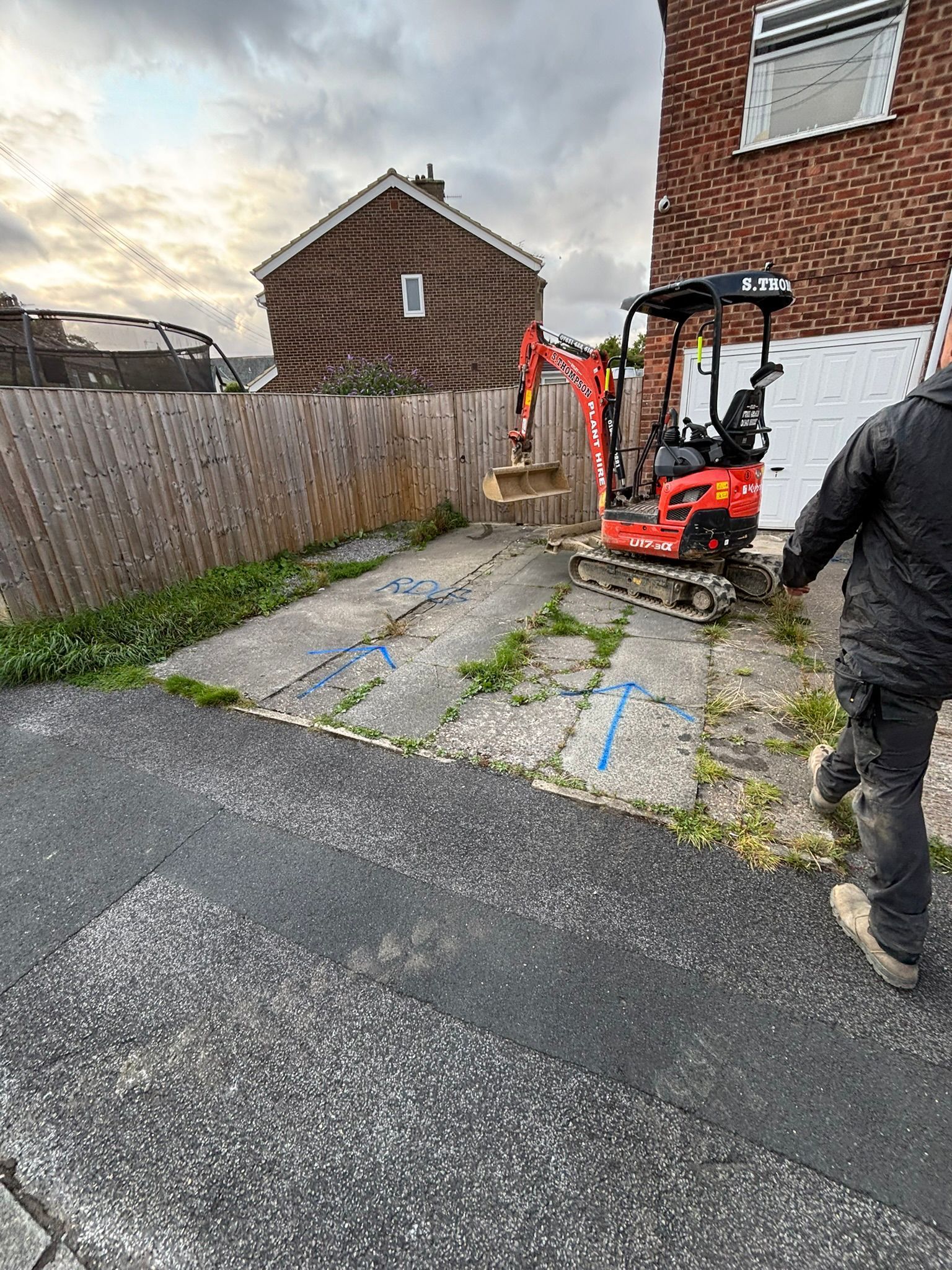 Worker beside red excavator on a paved driveway next to a house and fence