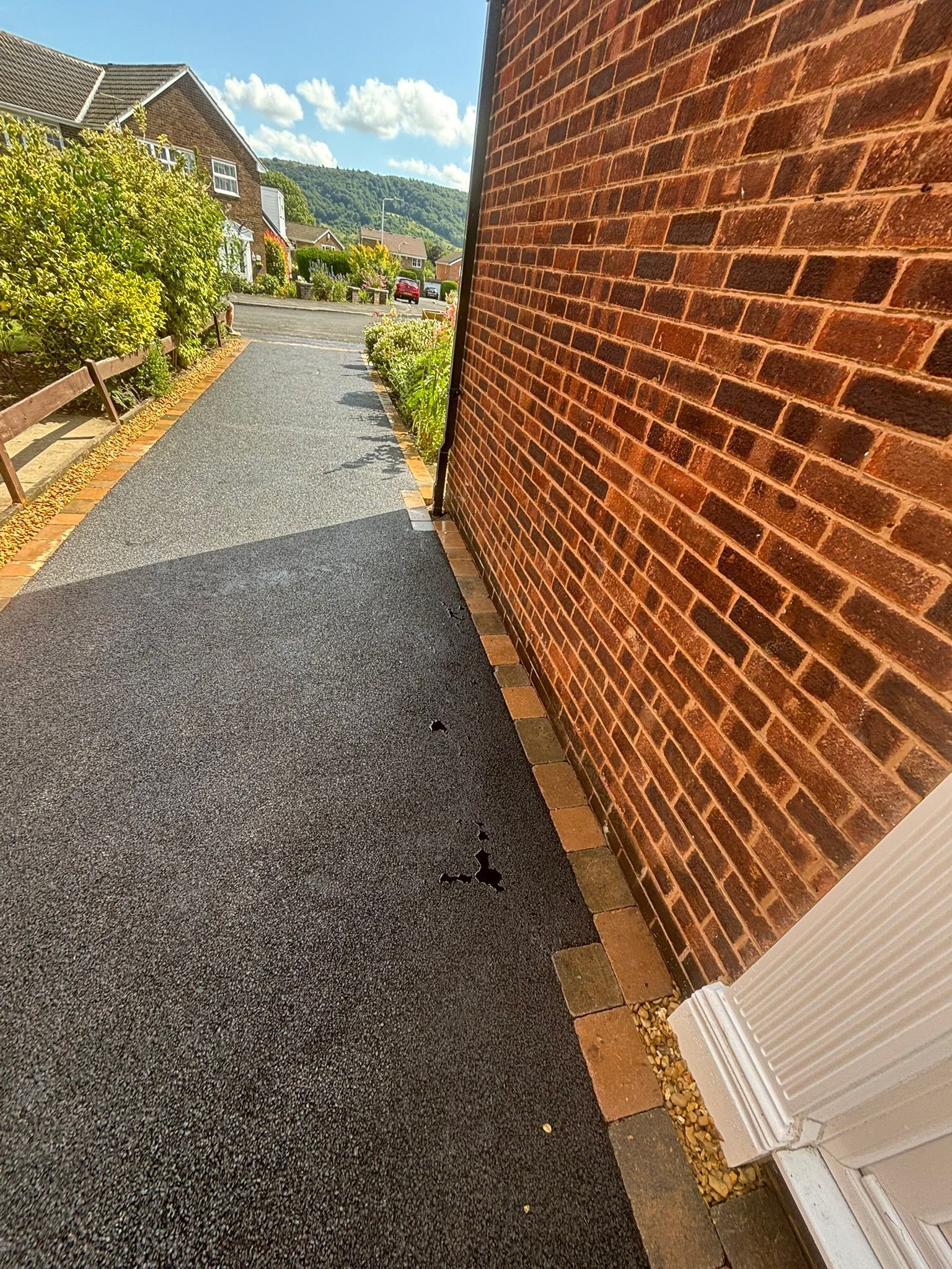 Narrow gravel path beside a red brick wall, leading to a sunny garden with mountains in the distance