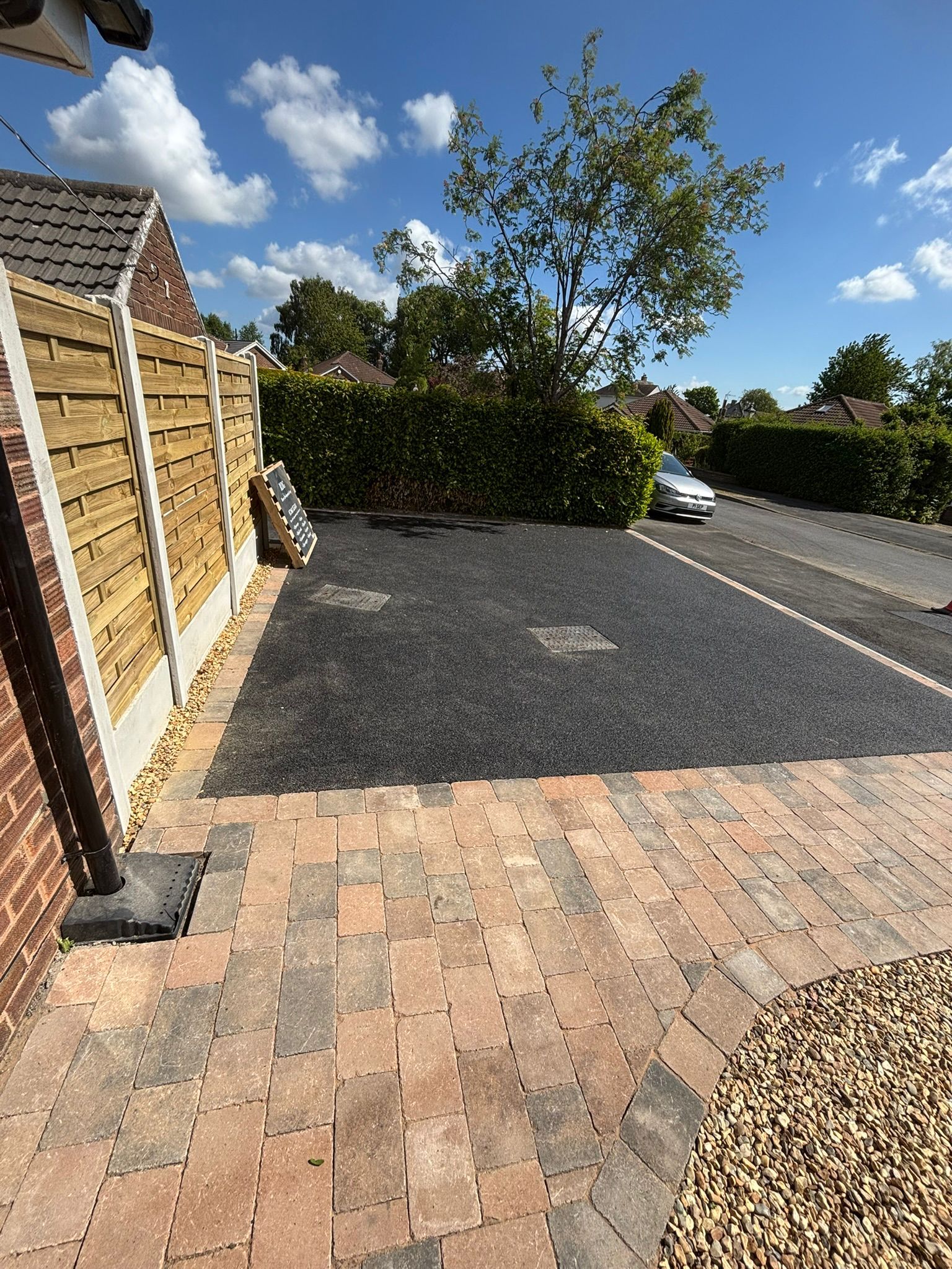 Brick driveway beside a wooden fence, gravel lot, and tree under a partly cloudy sky.