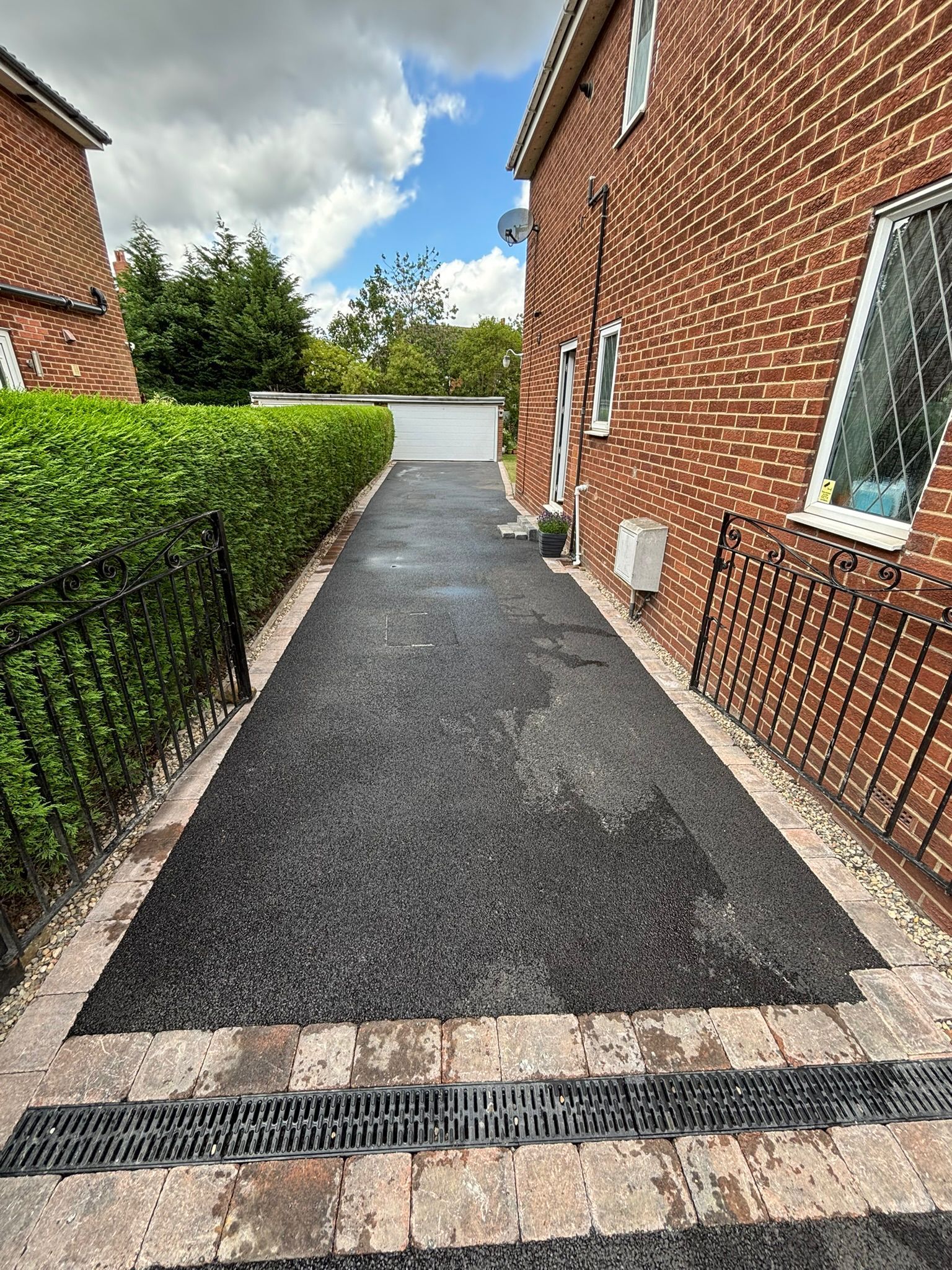 Narrow paved path beside red brick houses and hedges leading to a driveway under a cloudy sky
