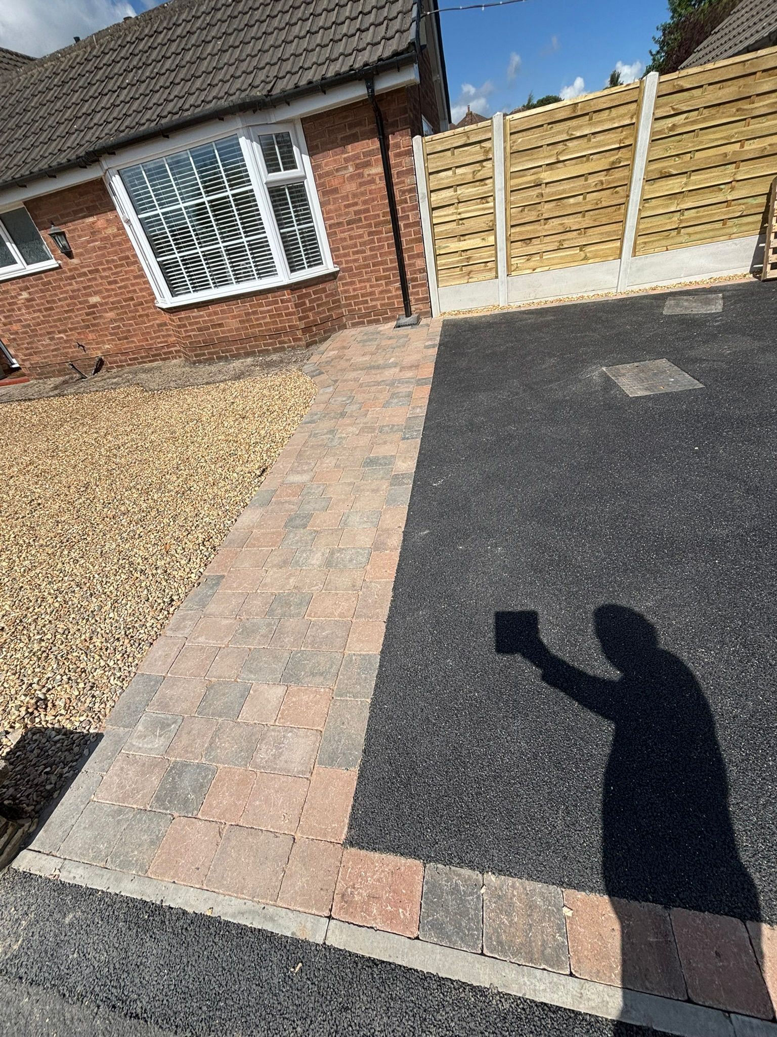 Brick and gravel driveway beside a house, with a black paved section and a fence gate.