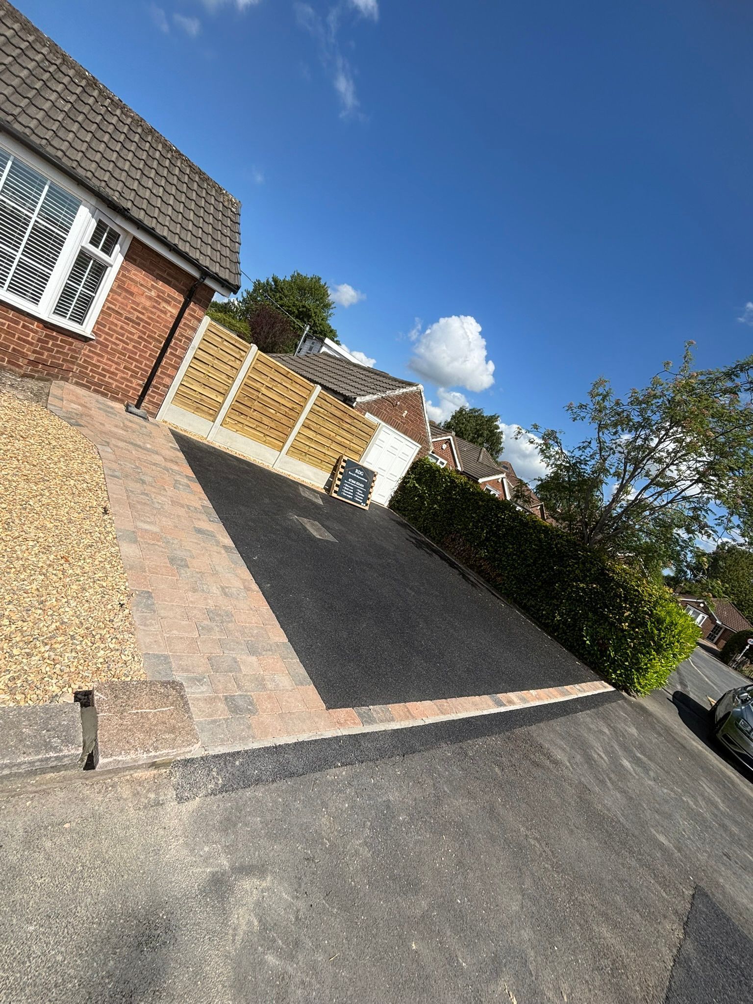 Tilted view of a suburban driveway with brick house, gravel and paved surfaces, wooden gate, and blue sky
