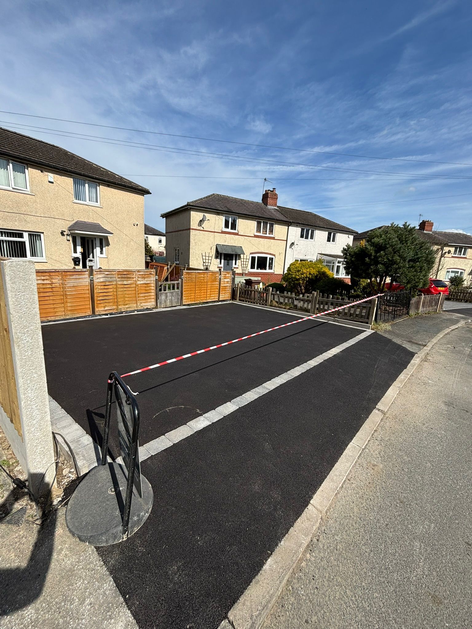 Residential street with fresh black road resurfacing, red-and-white barriers, and houses under a blue sky