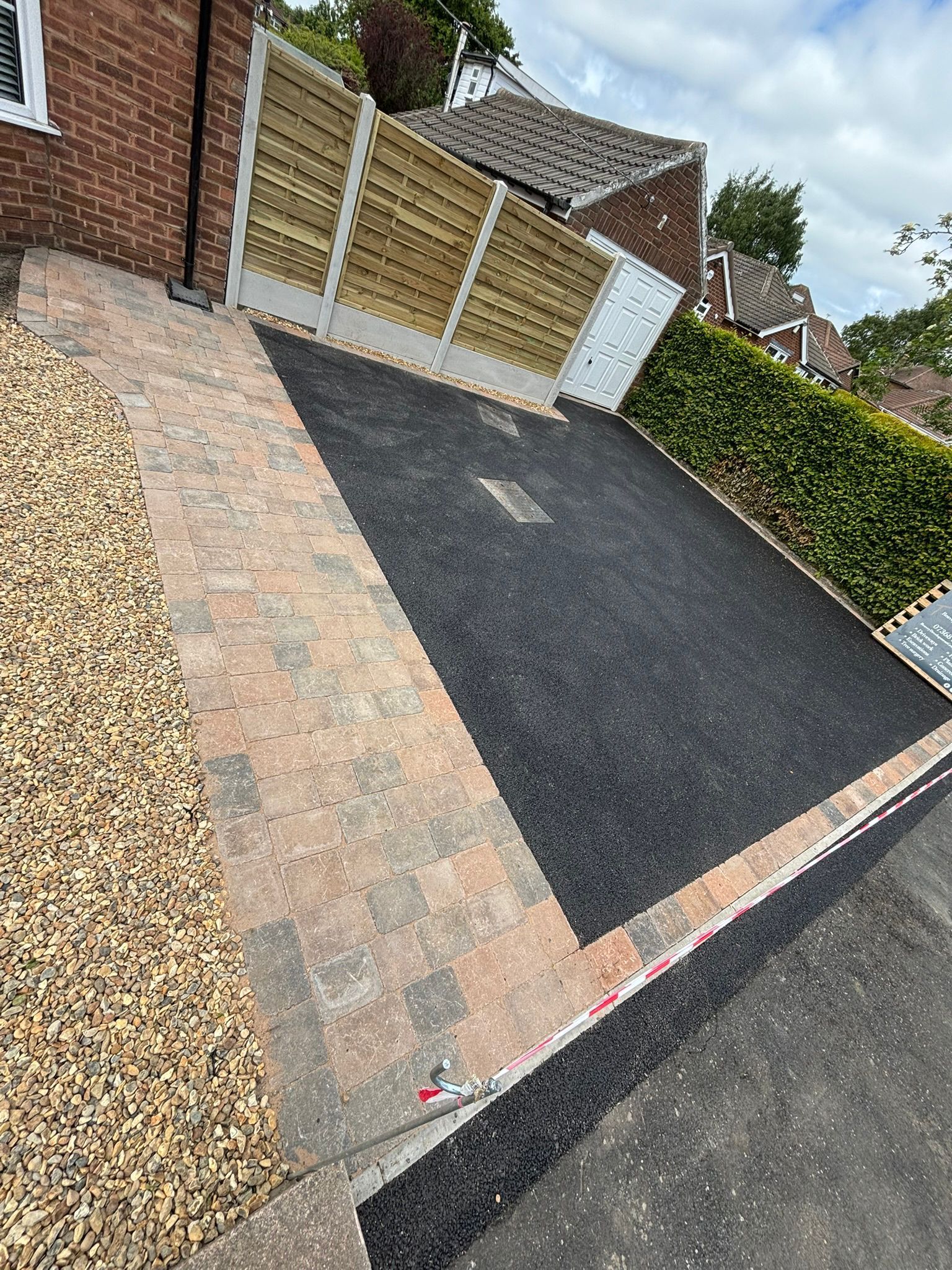 Tilted view of a paved driveway beside a brick house and hedges, with a gravel strip on the left.