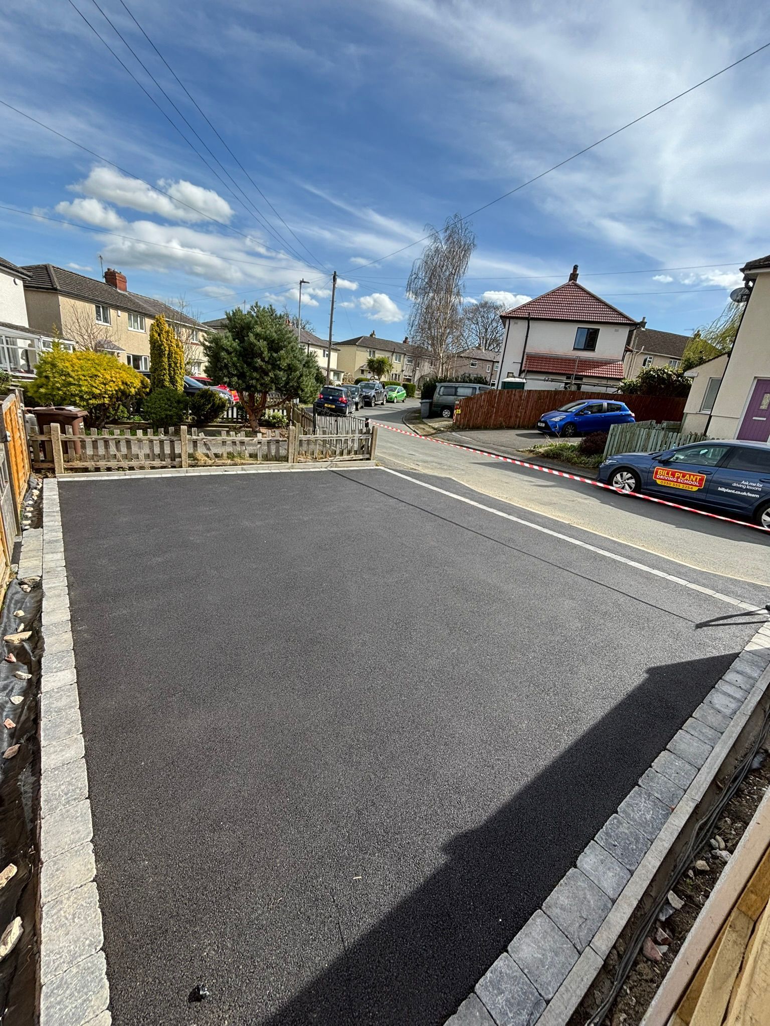 Quiet residential street with freshly paved road, houses, parked cars, and a bright blue sky