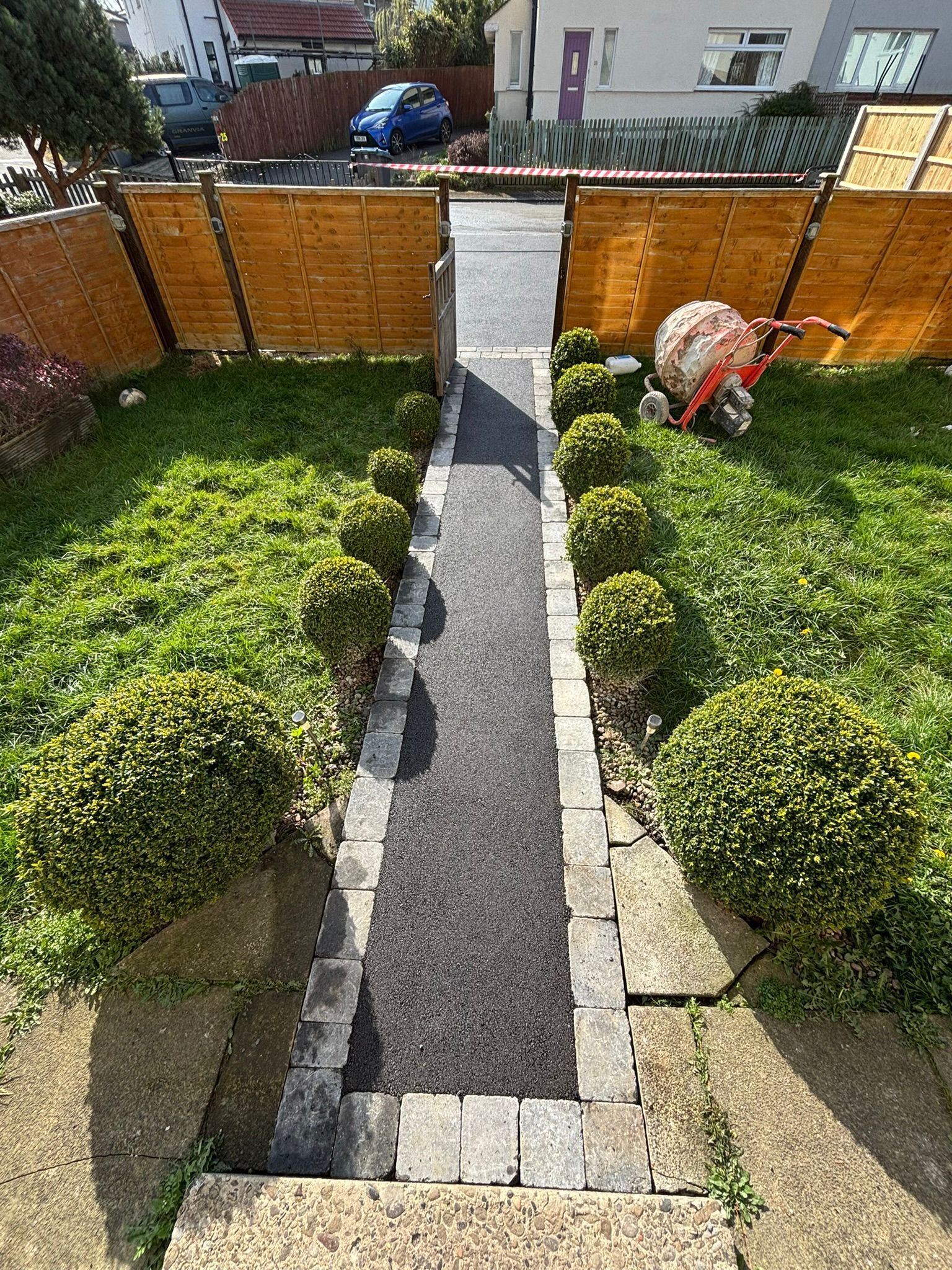 Front garden path between trimmed hedges leading to a wooden gate, with a gardener working on the right.