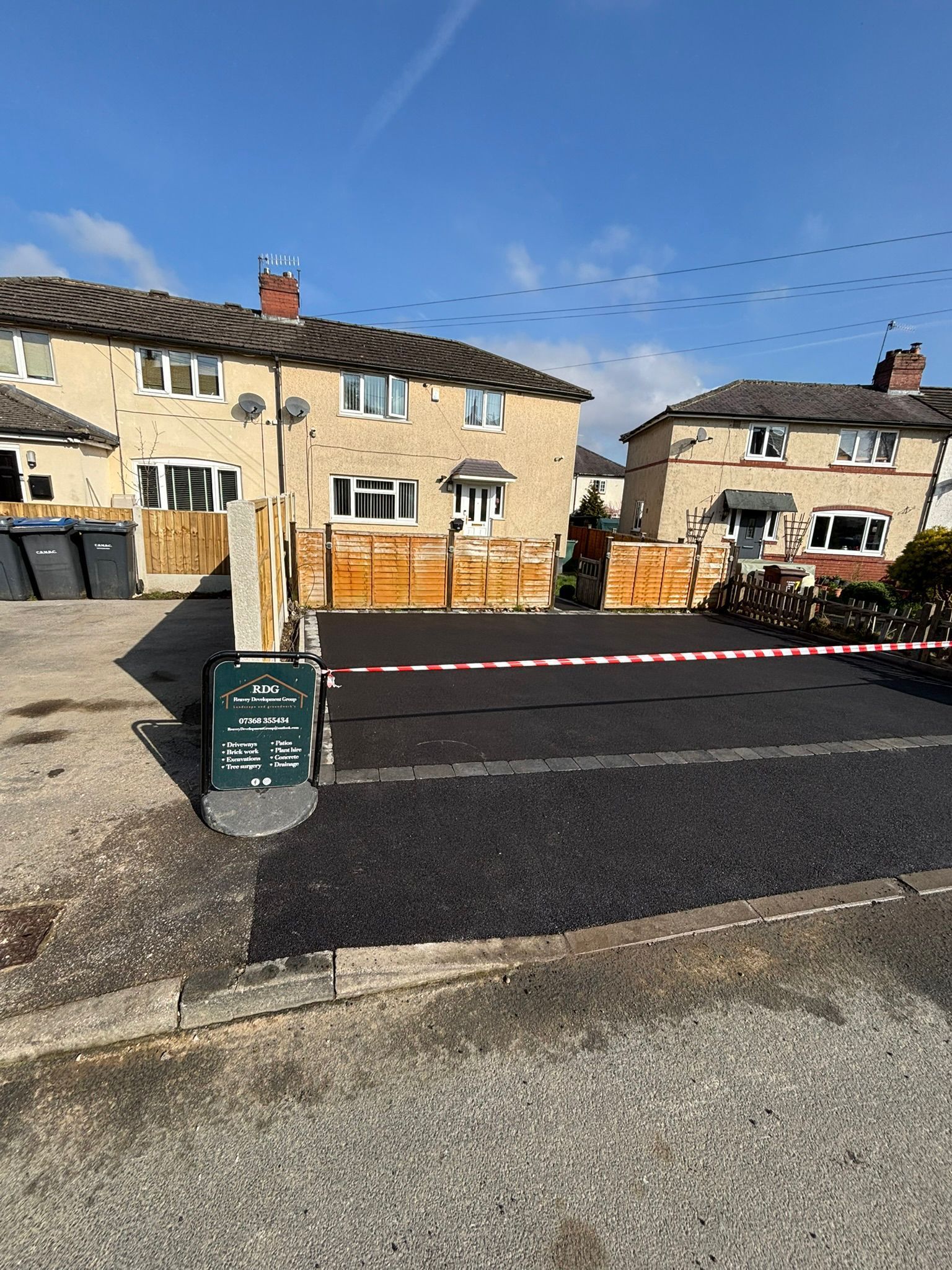 Residential street with stone houses, road closed by red-and-white barrier, and a black trash bin in the foreground