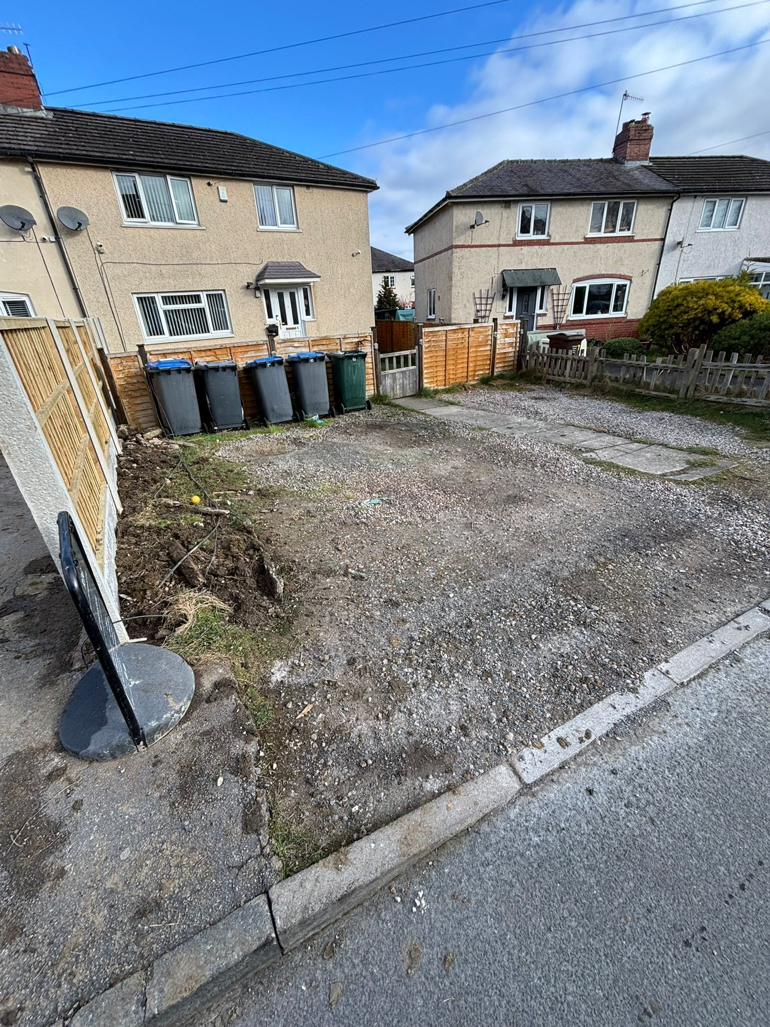 Gravel driveway beside two beige houses with trash bins and a wooden gate under a blue sky