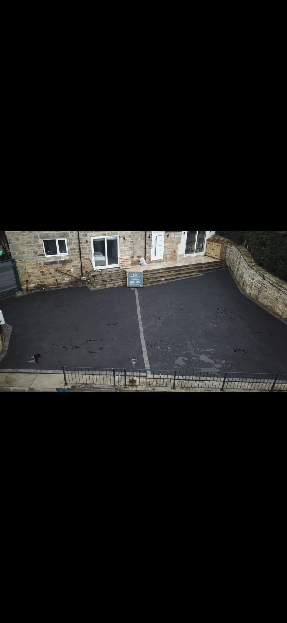 Freshly paved black driveway in front of a stone house, with a wheelbarrow and tools near the entrance.