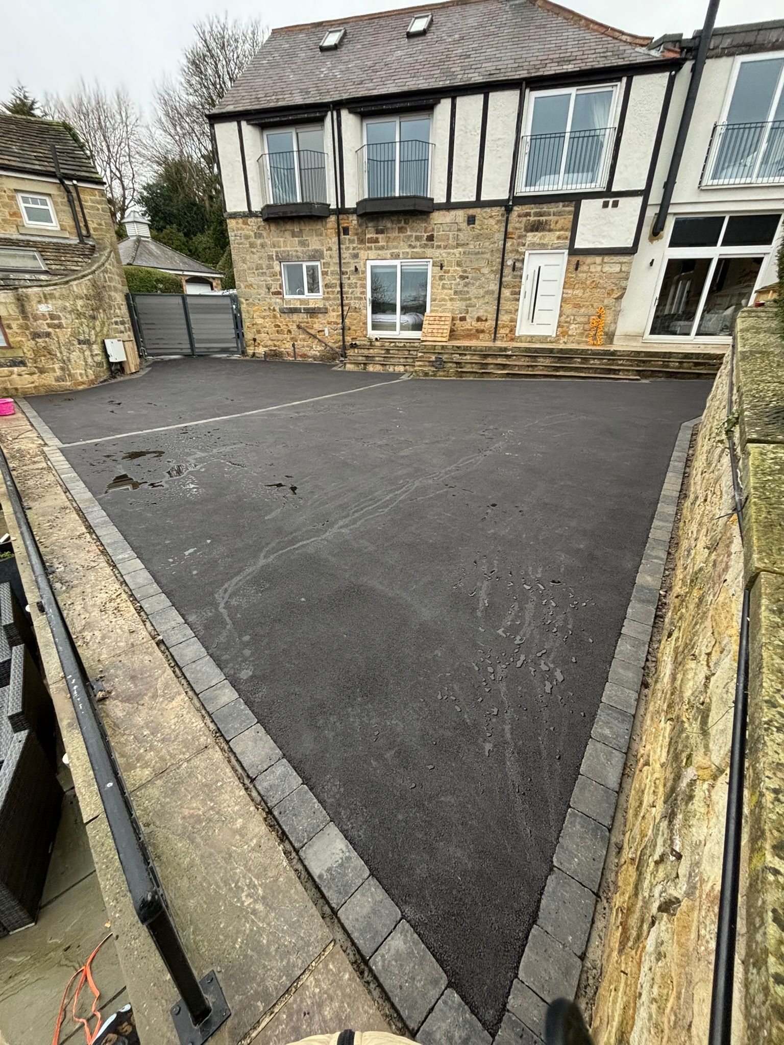 Stone driveway with fresh black asphalt leading to a house with a bay window and stone walls