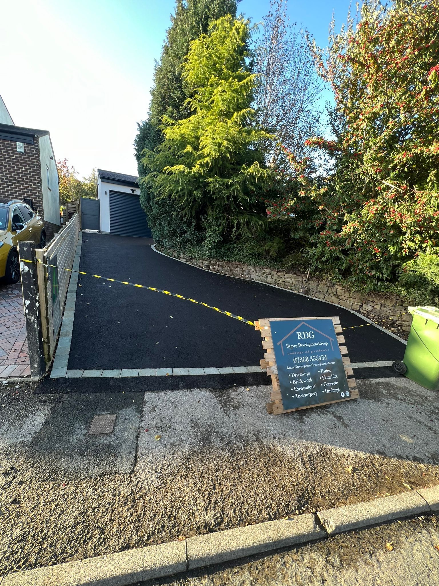 A driveway with a rope barrier, a signboard, and tall trees beside a house and garage.