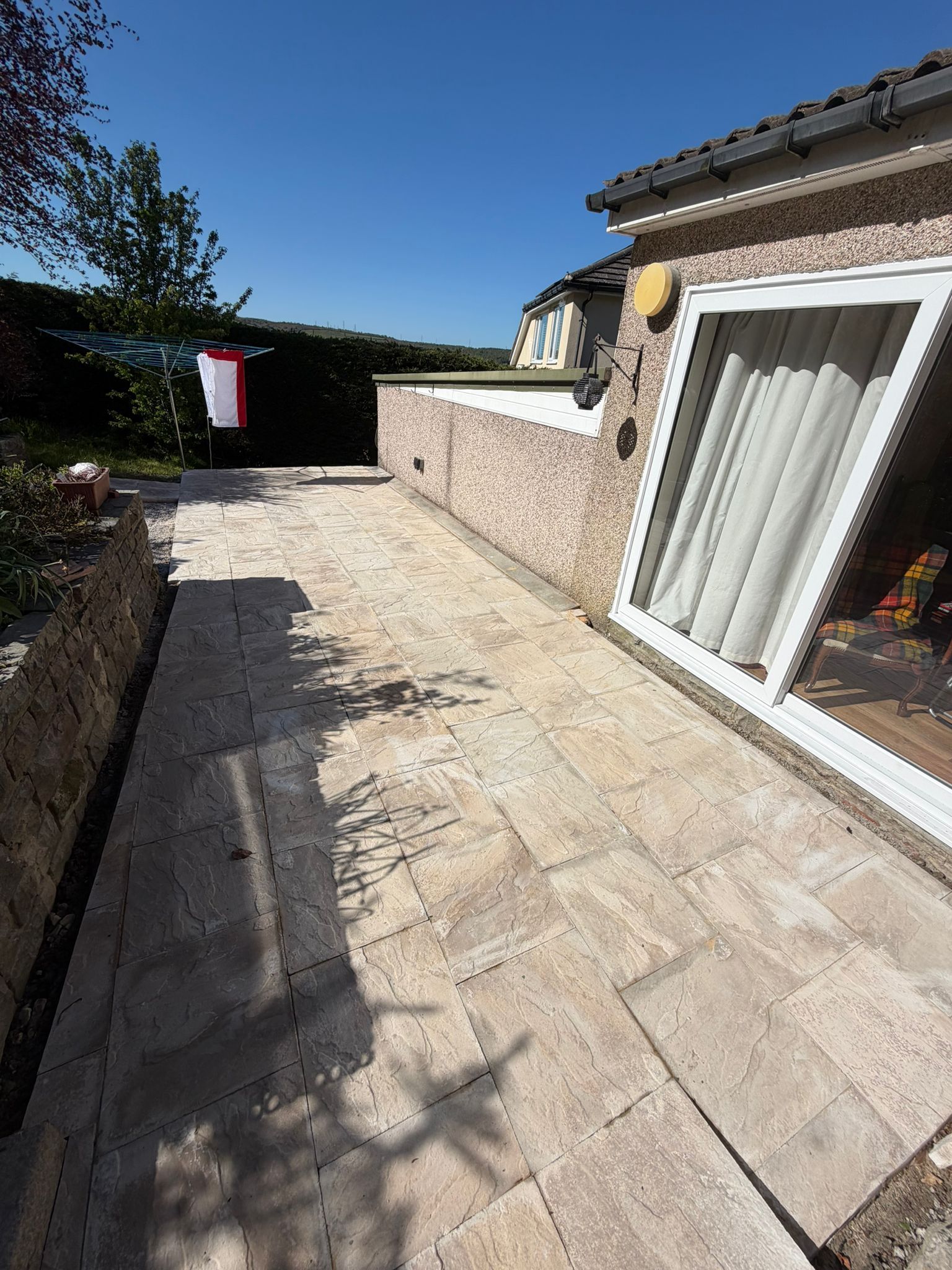 Sunlit stone patio beside a house, with hedge, wall, and hanging laundry in the background.