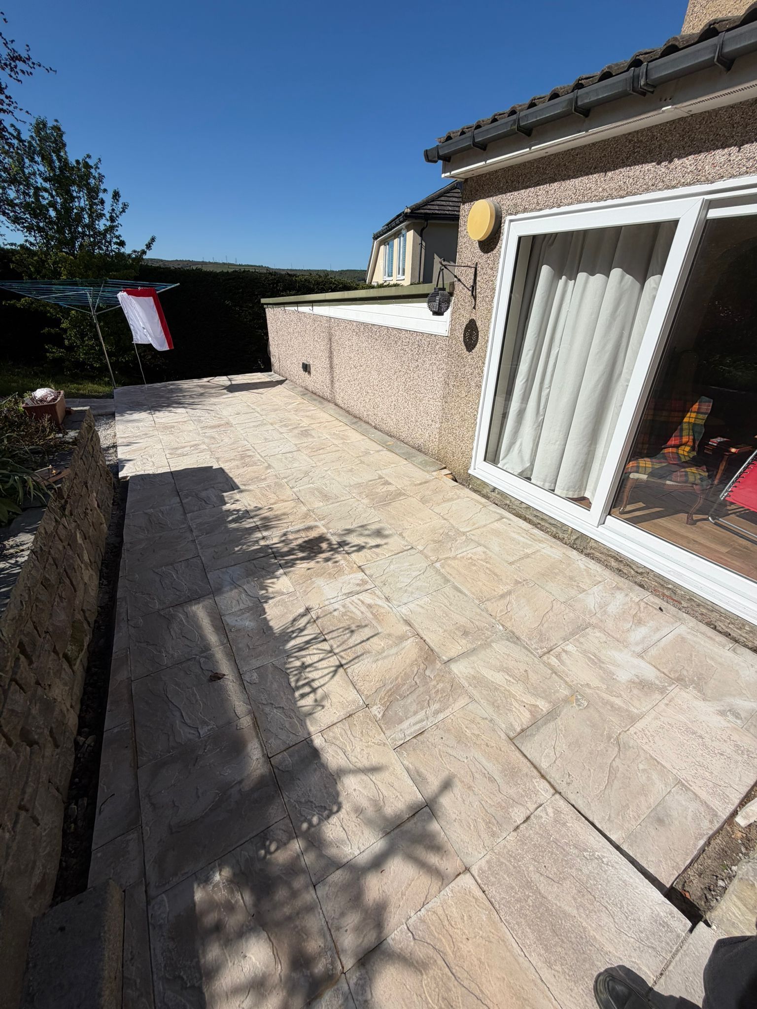 Sunlit patio with stone paving beside a house, bordered by a wall and hedge, with a sliding glass door open.
