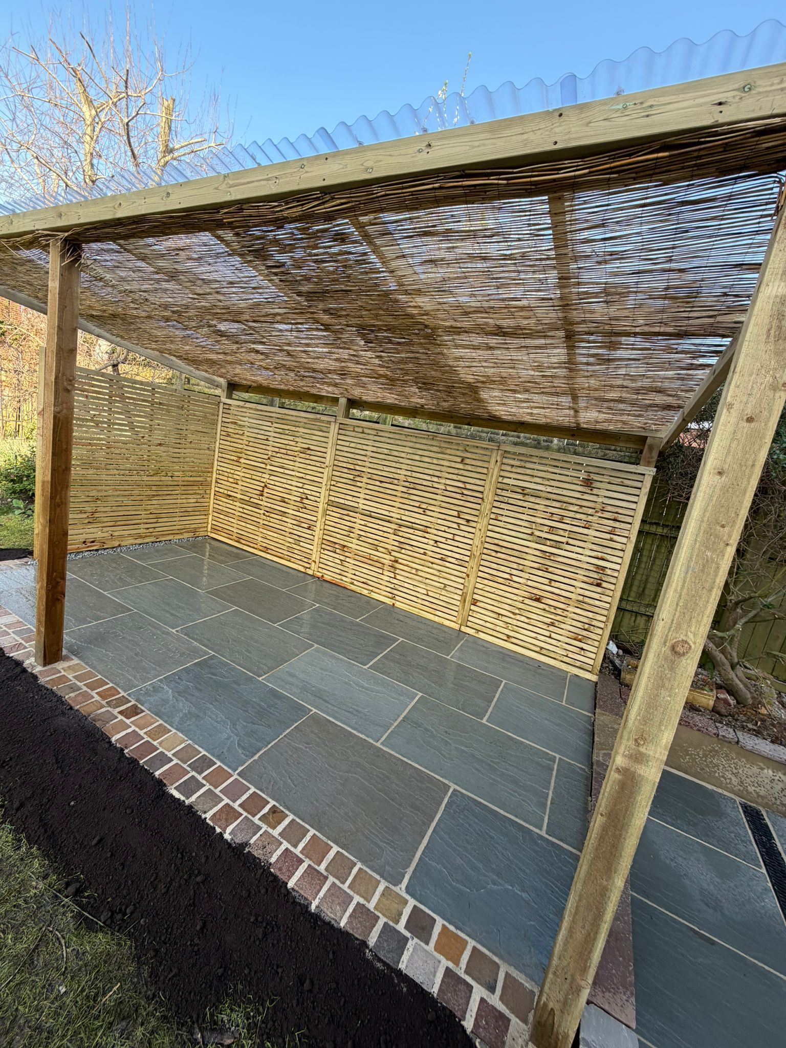 Outdoor patio with woven wood canopy, patterned wall, and dark stone flooring in a sunlit garden area